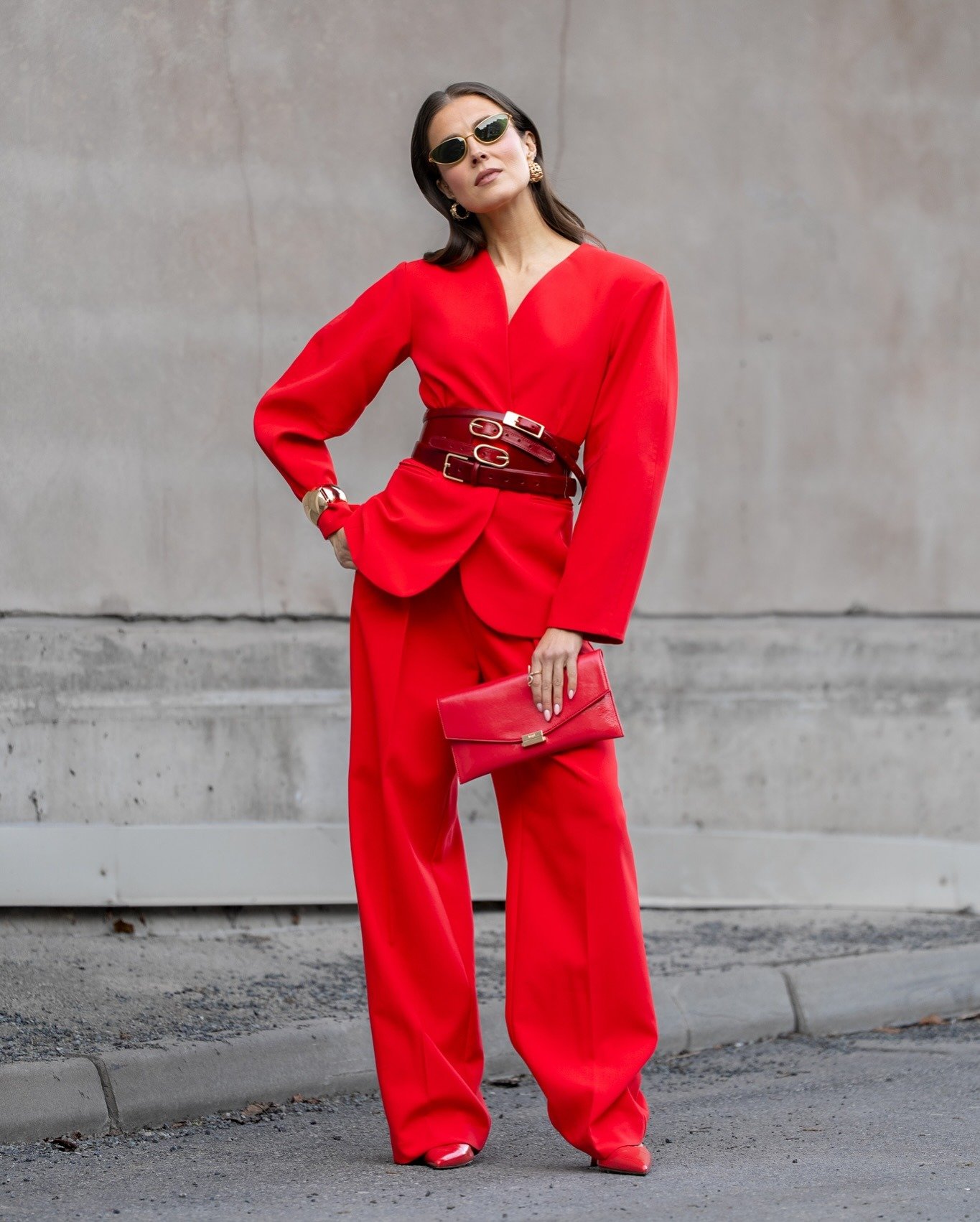 Este look colorido para trabalhar destaca um conjunto vermelho vibrante, composto por um blazer de mangas largas e calças amplas. A cintura é acentuada por um cinto largo em tom semelhante, adicionando sofisticação. Acessórios como óculos escuros e uma cl