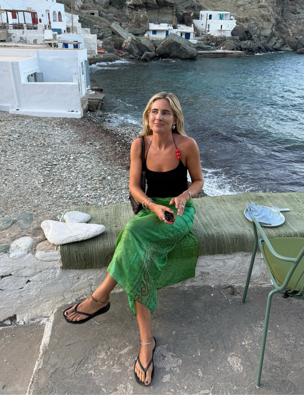 Mulher sentada à beira-mar com saia verde e regata preta, criando looks frescos no cenário litorâneo.