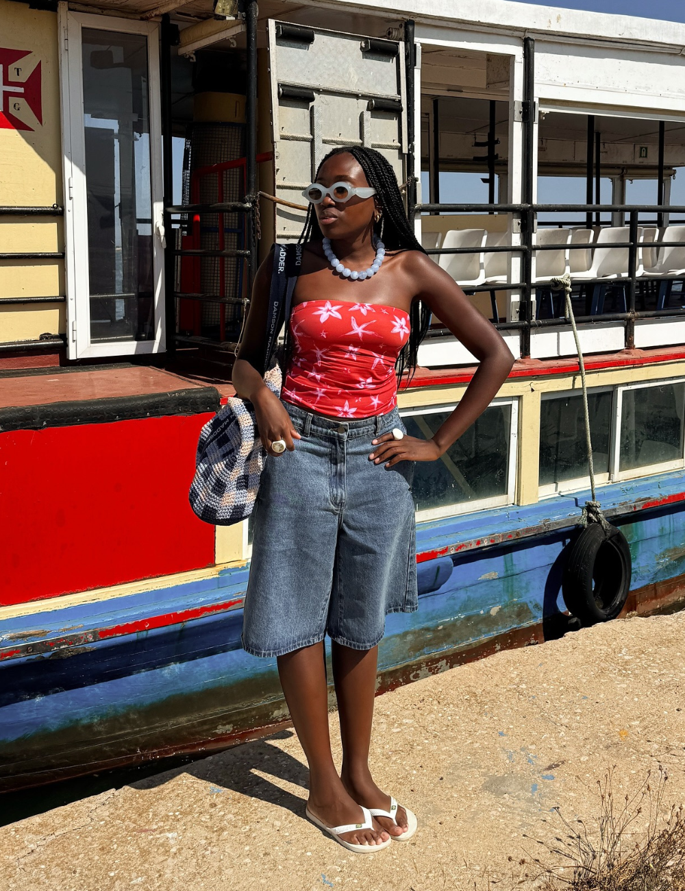 Usuária com blusa vermelha sem alças e motivos florais, short jeans e óculos, em frente a um barco.