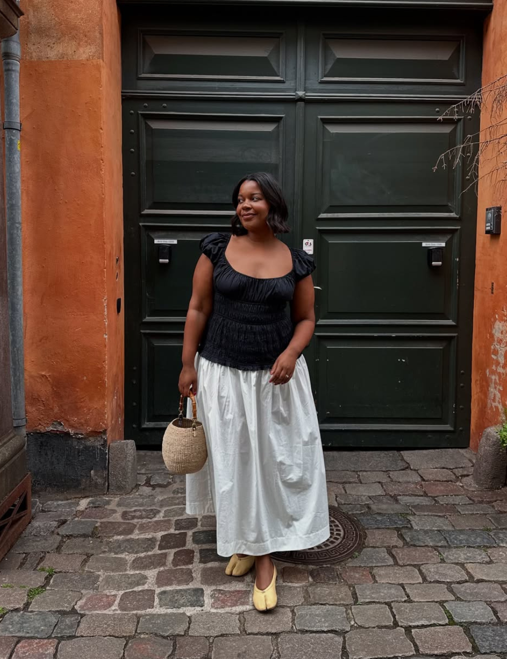 Pessoa em frente a um portão, vestindo blusa preta com saia longa branca. Look casual com blusa fresca.