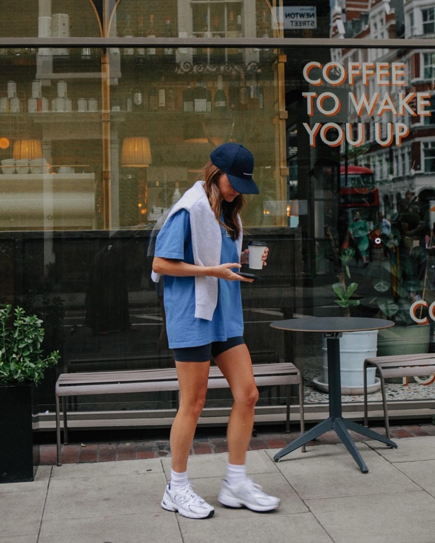 Mulher com boné feminino azul, camiseta larga com toalha nos ombros e tênis, passa em frente a uma cafeteria.
