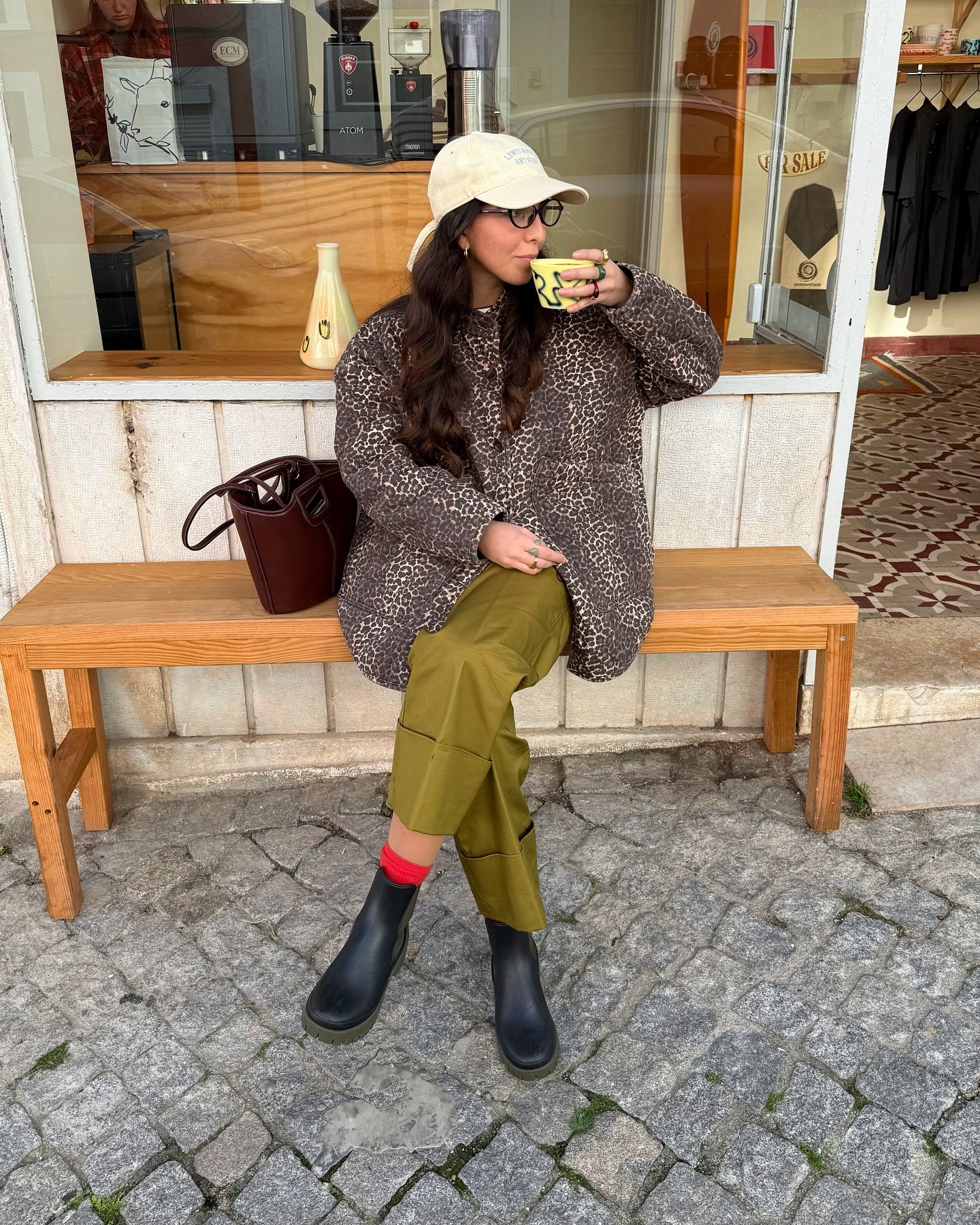 Mulher com casaco de estampa animal e boné feminino, sentada em banco, tomando café.