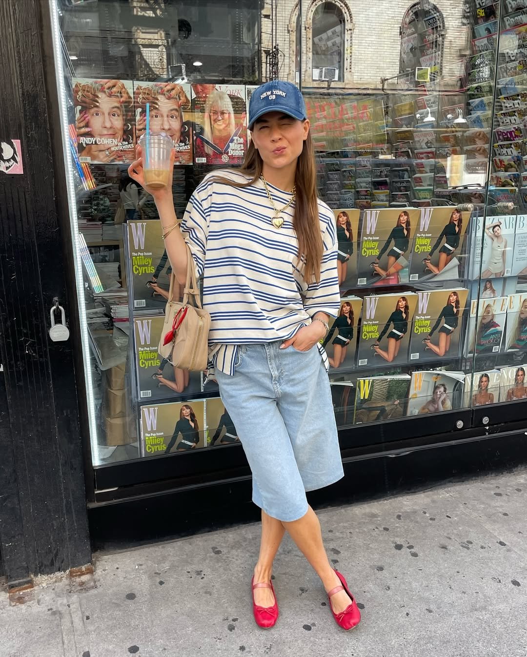 Pessoa com boné azul, blusa listrada, bermuda jeans e sapatilhas rosas em frente a banca de revistas.