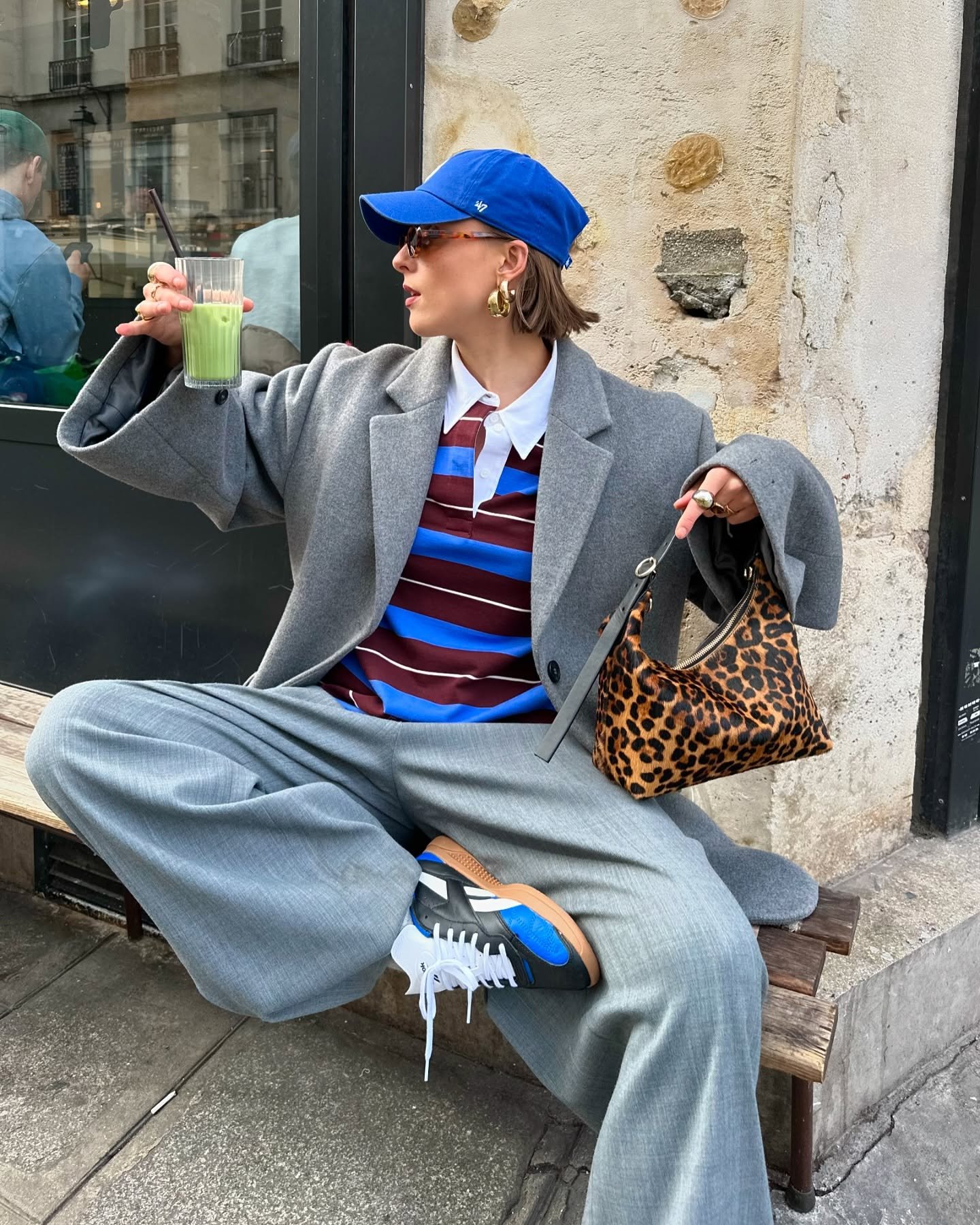 Pessoa com boné azul, casaco cinza e bolsa estampada, segurando um copo de suco. Representando looks com boné feminino.