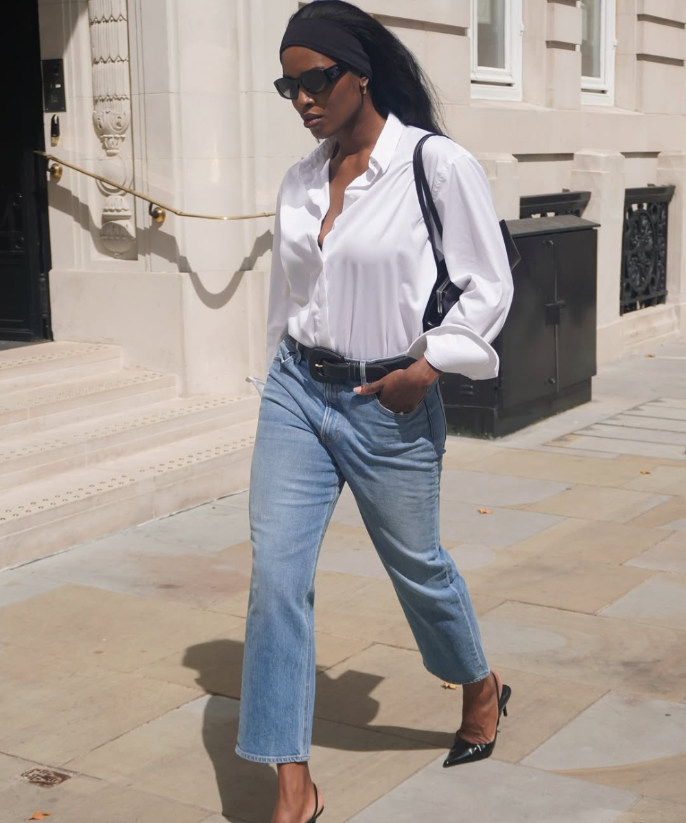 Mulher caminhando com calça jeans e salto alto, usando camisa branca e óculos escuros.