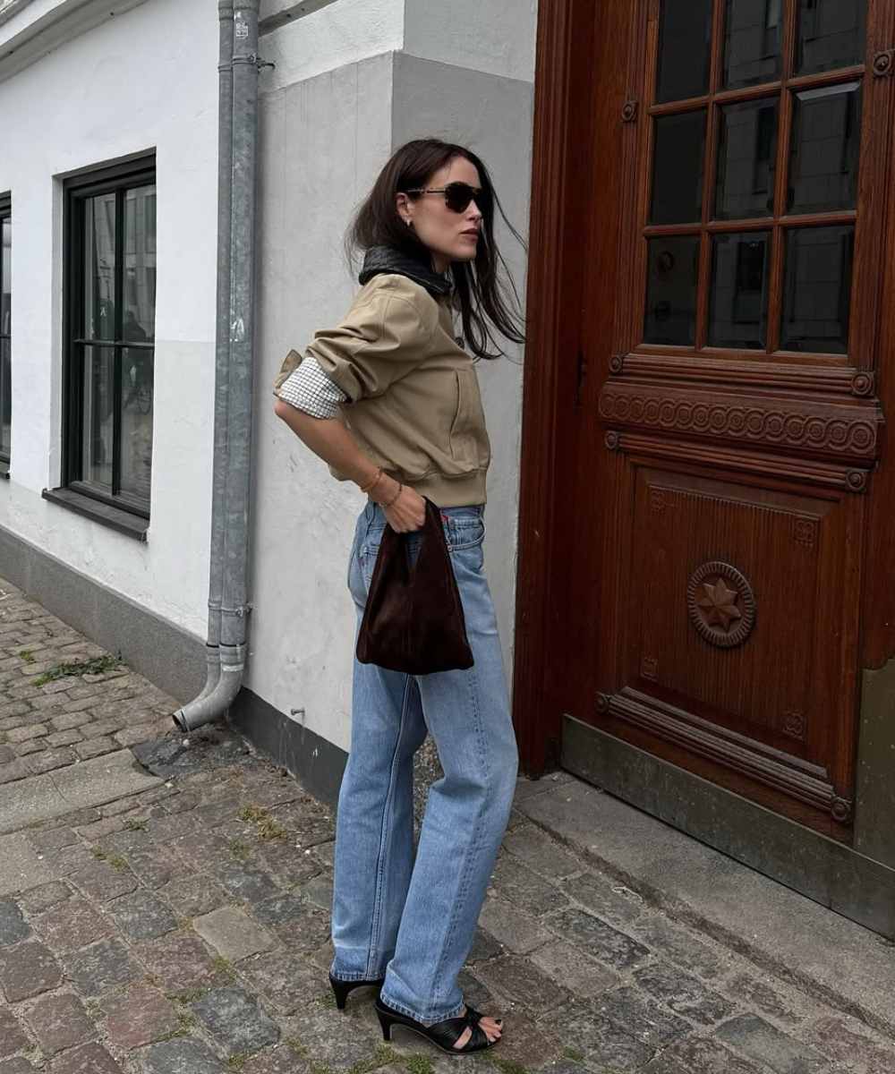 Pessoa usando jeans e salto alto, segurando bolsa marrom em frente a uma porta de madeira.