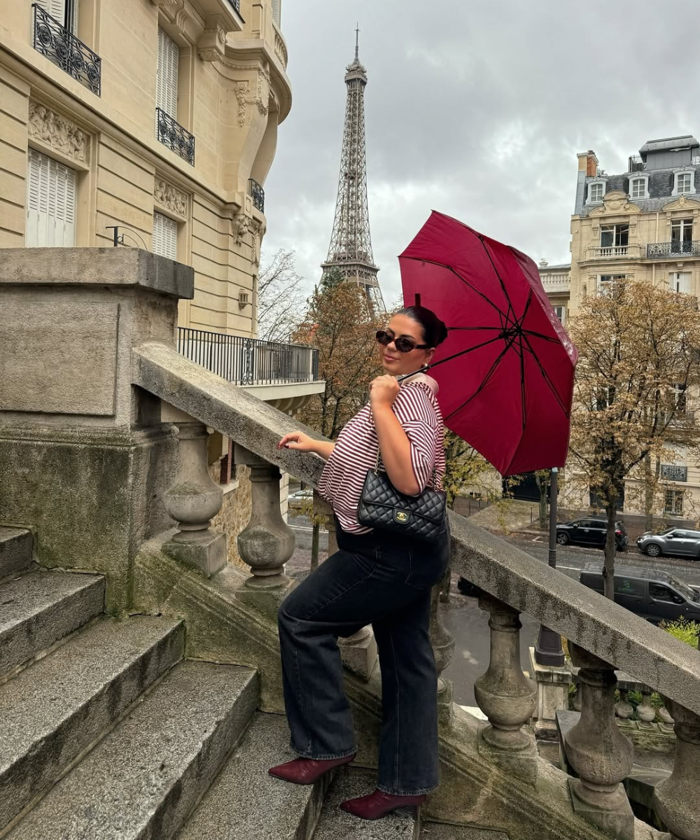 Pessoa em escadaria vestindo calça jeans e salto alto, segurando guarda-chuva vermelho com Torre Eiffel ao fundo.