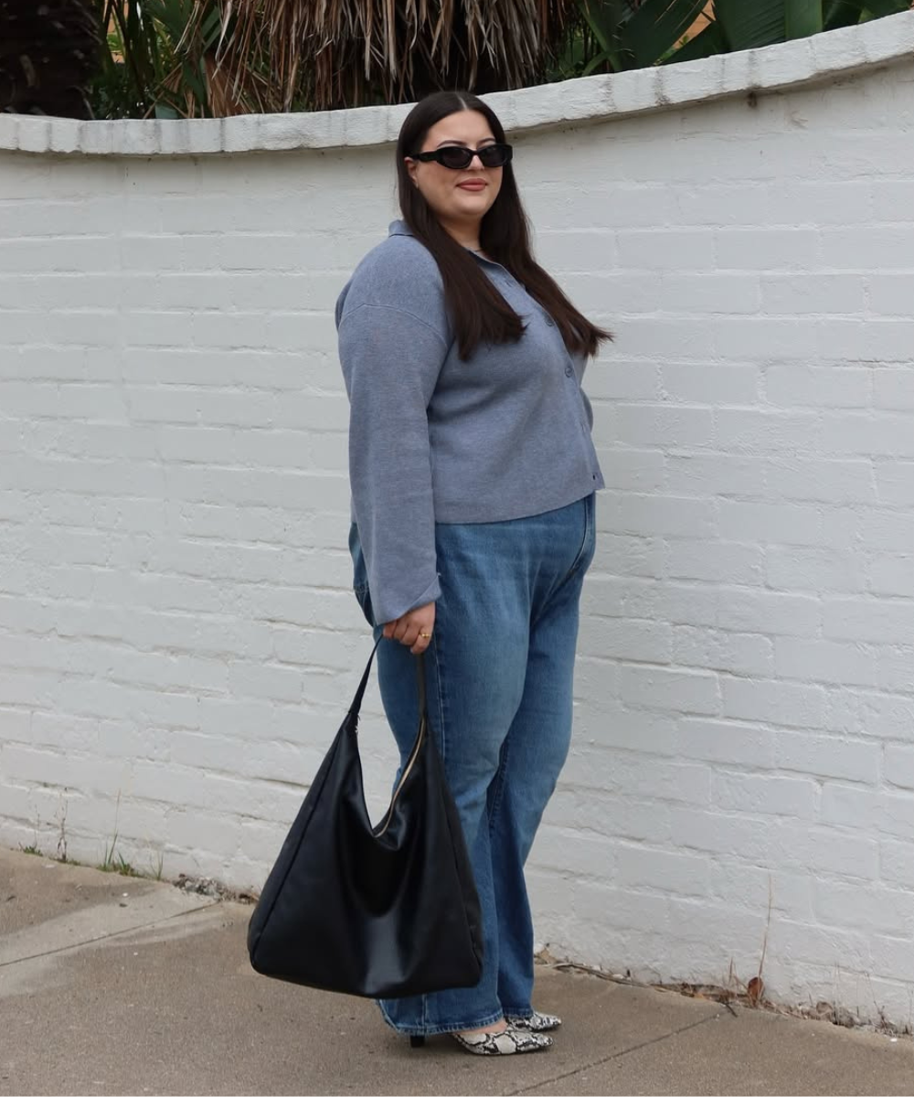 Mulher usando suéter cinza com calça jeans, salto alto estampado e bolsa preta, perto de muro branco.