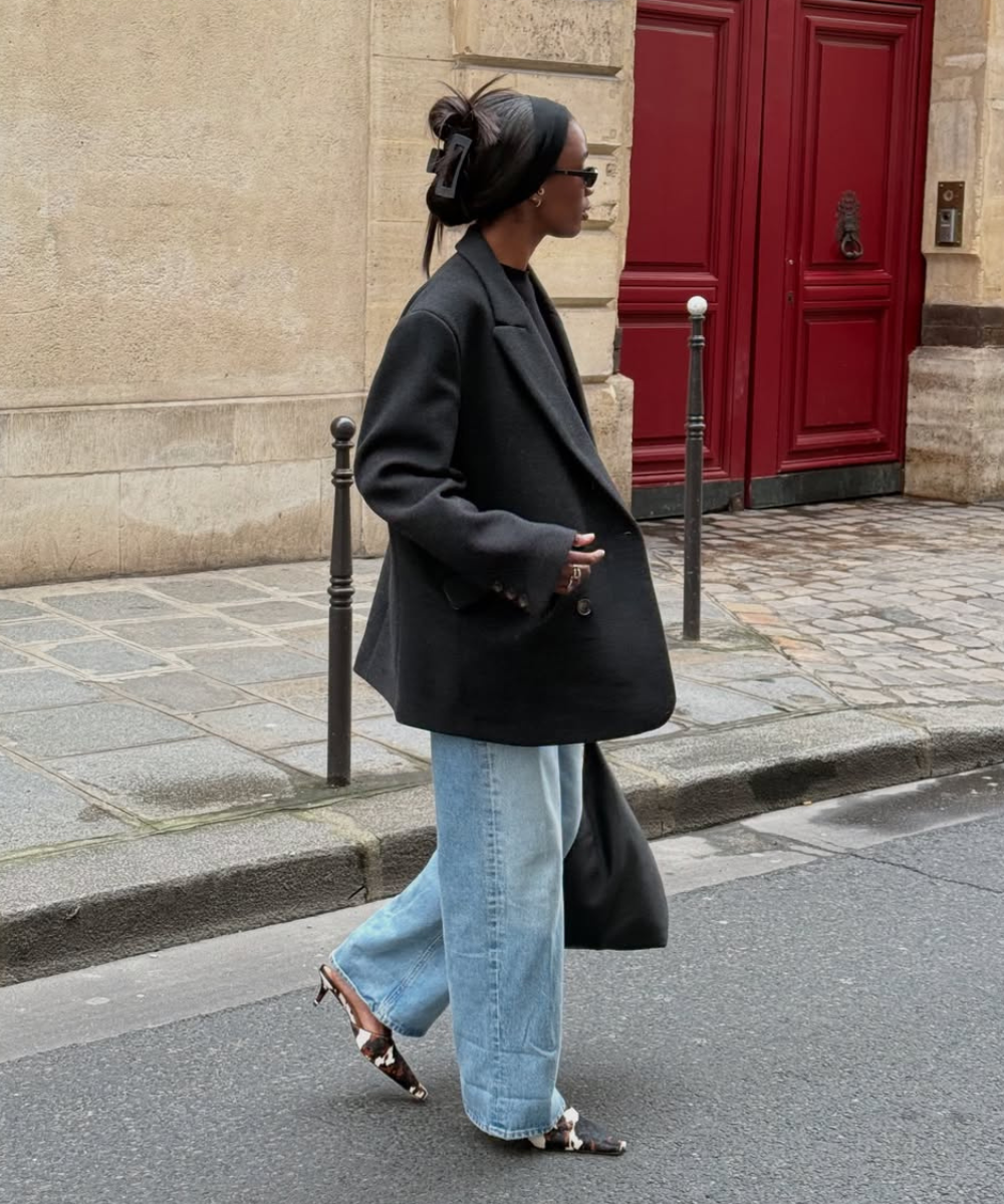 Pessoa usando casaco preto, calça jeans larga e salto alto estampado, caminhando em rua de paralelepípedos.