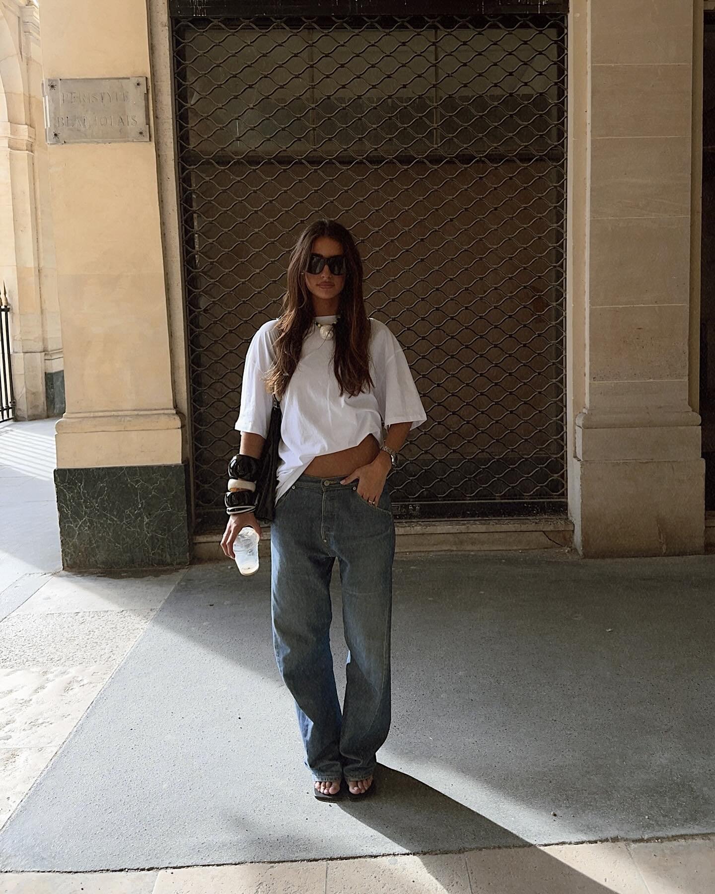 Mulher em look casual com calça jeans e camiseta branca, segurando garrafa e usando óculos escuros.