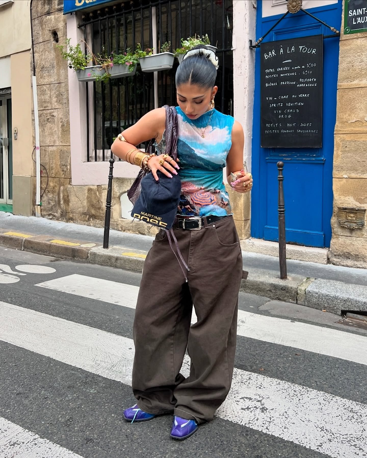 Pessoa em pé na rua, usando calça marrom feminina, top colorido e segurando uma bolsa escura.