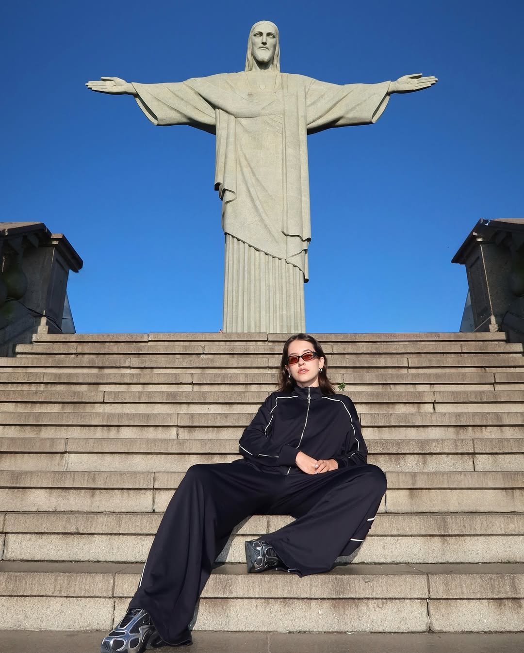 Mulher com look esportivo usando calça preta larga e jaqueta combina tênis modernos em frente ao Cristo Redentor.
