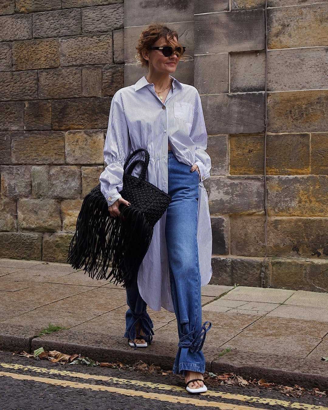 Mulher usa chemise listrada longa, jeans e bolsa de franjas negras em frente a parede de pedra.