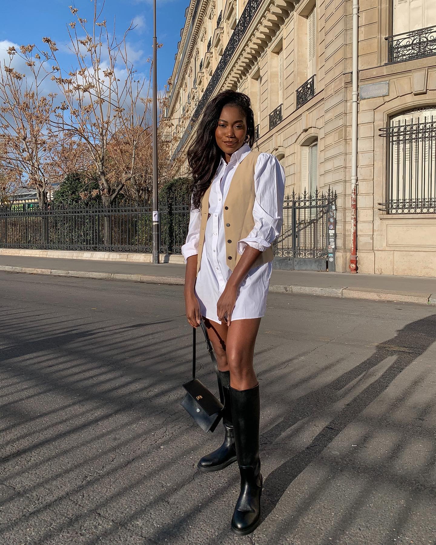 Pessoa em pé na rua usando chemise branca com colete bege, botas pretas e bolsa.