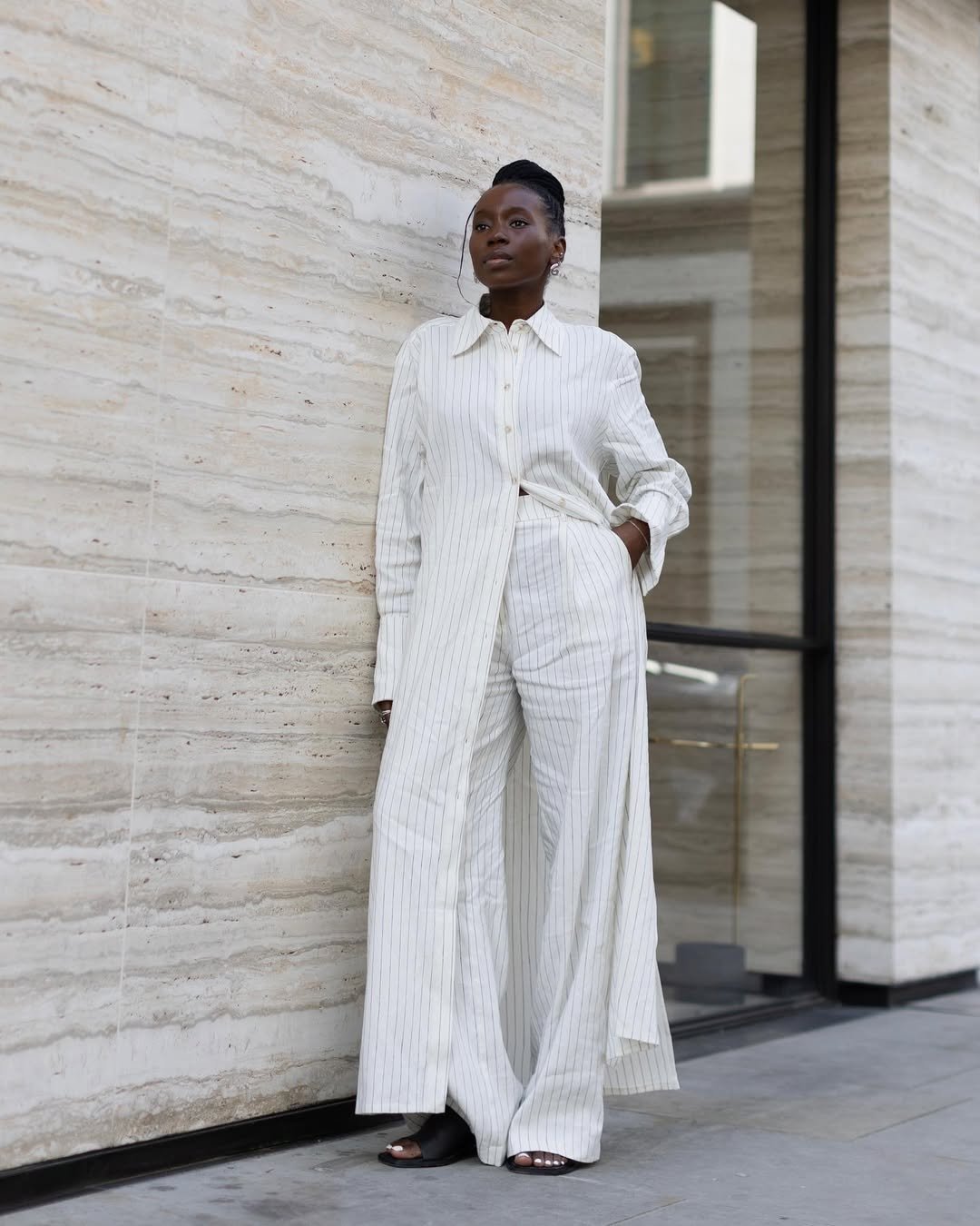 Pessoa em traje elegante com chemise e calça listradas, encostada em parede marmorizada.