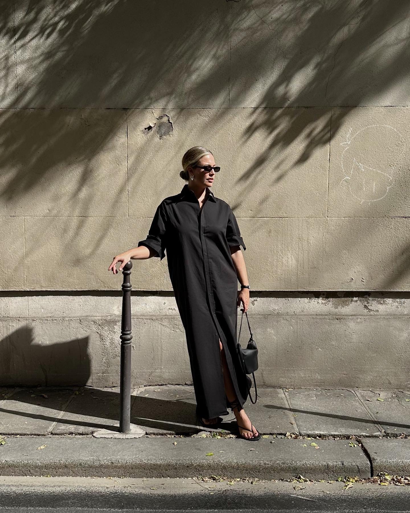 Pessoa usando chemise preta em rua ensolarada, segurando bolsa pequena e usando óculos escuros.