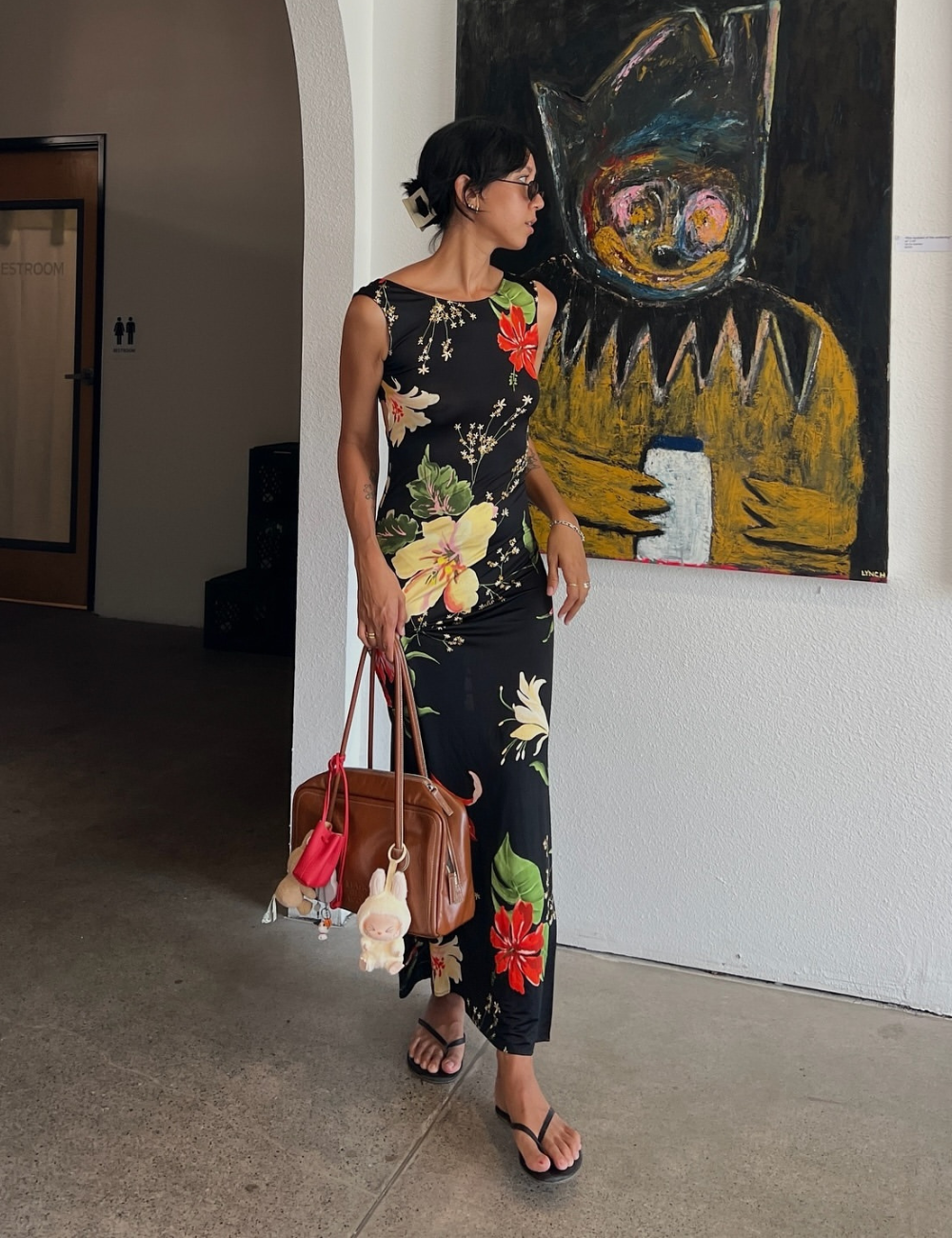 Mulher com vestido floral preto e chinelos, segurando bolsa marrom, em frente a uma pintura colorida na galeria.