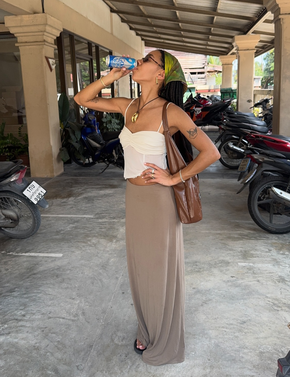 Pessoa bebe água de coco, usando saia longa e chinelo preto, em um estacionamento.