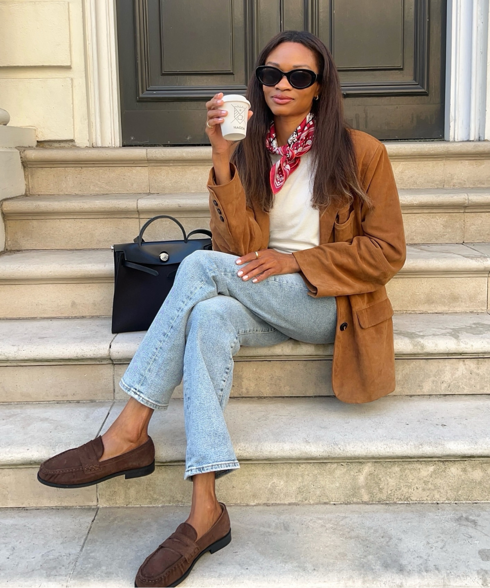 O mocassim de camurça é destacado neste look elegante e descontraído. A modelo, sentada em escadas, combina o calçado com jeans claros e uma blusa básica, complementando com um blazer de camurça. Um lenço colorido no pescoço e óculos escuros adicionam sof