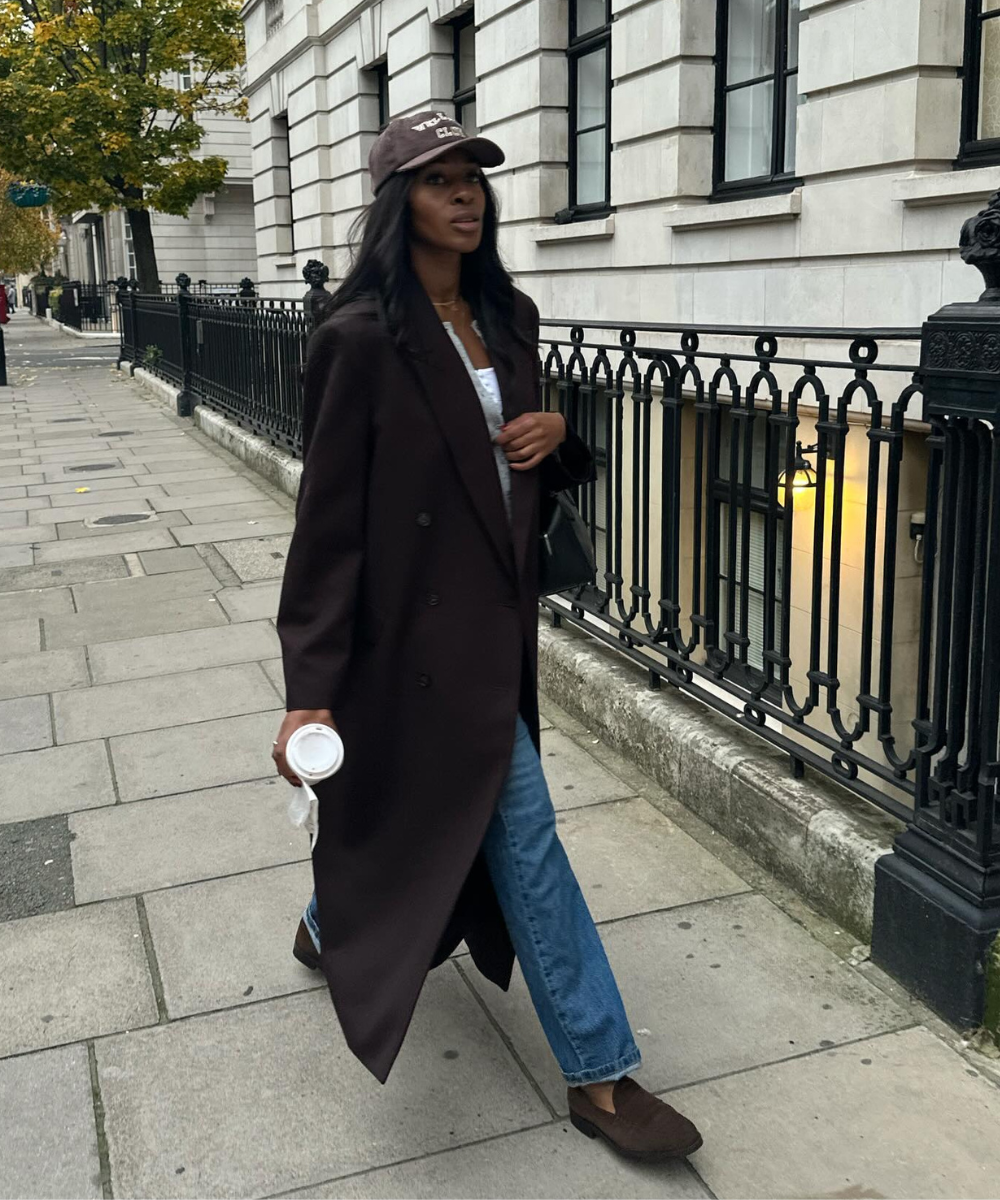 A imagem apresenta uma mulher caminhando em uma calçada, vestindo um longo casaco marrom e jeans claros. Nos pés, um elegante mocassim de camurça marrom, que complementa o visual casual e sofisticado. O look é finalizado com um boné e acessórios discretos