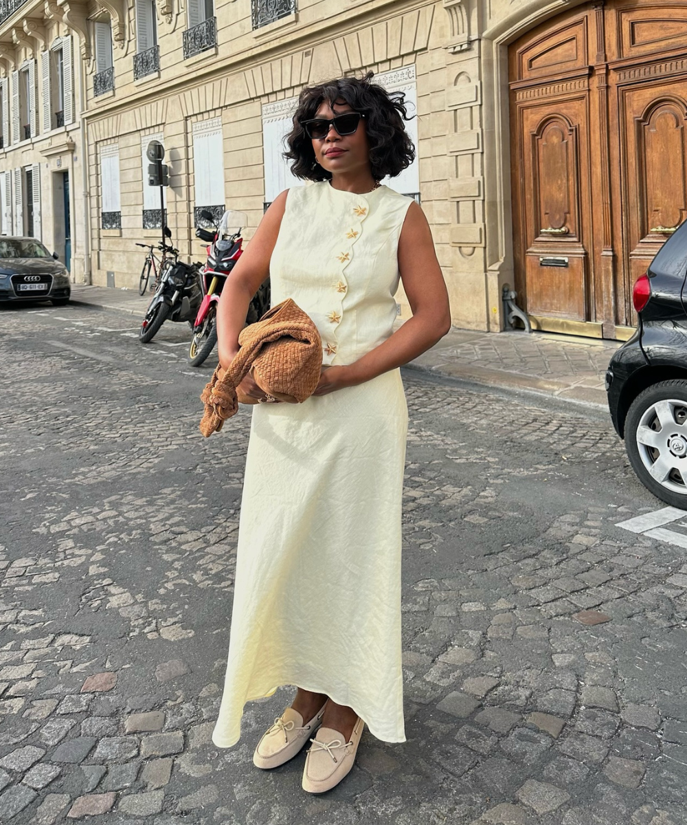O mocassim de camurça destaca-se no look elegante, complementando um vestido amarelo claro com detalhes florais. Os óculos escuros e a bolsa de palha adicionam um toque moderno e descontraído. A combinação é perfeita para um passeio urbano, refletindo sof