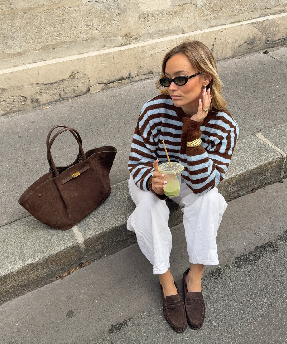 O mocassim de camurça, em tom marrom escuro, complementa o look casual e elegante da modelo. Ela usa um suéter listrado em azul e marrom, combinado com calças brancas. Acessórios como óculos escuros e uma bolsa de camurça adicionam sofisticação.