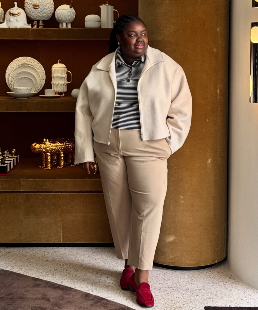O mocassim de camurça, em tom vermelho vibrante, complementa o look sofisticado. A modelo veste uma jaqueta oversized em tom claro, sobre uma blusa listrada, e calças em tom neutro, criando um contraste elegante.