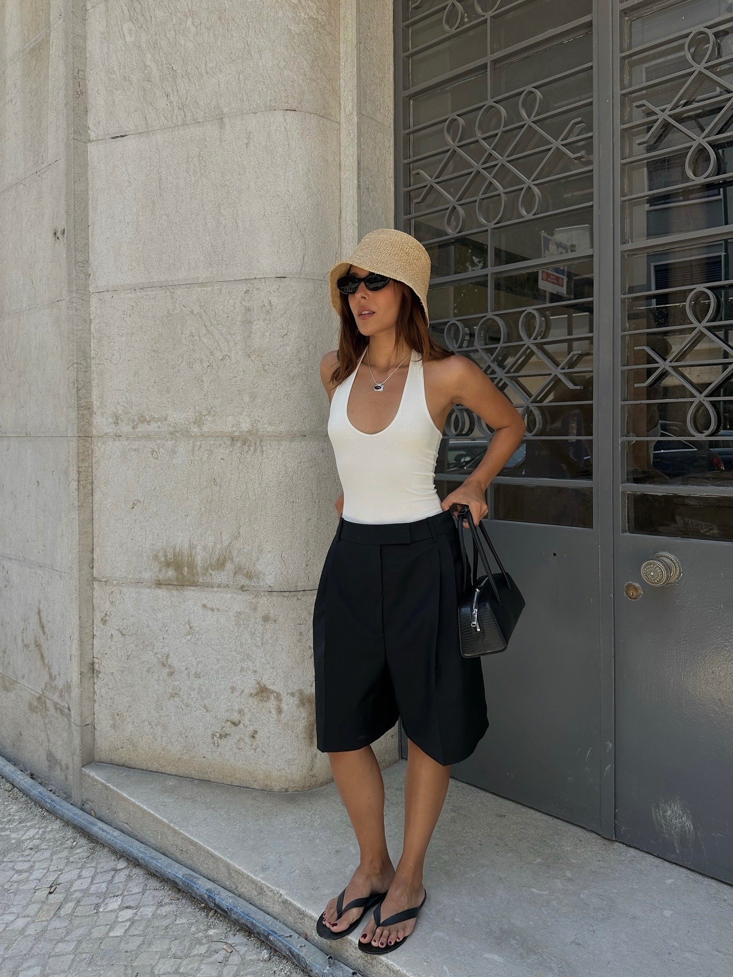 O look com regata apresenta uma combinação moderna e leve, ideal para dias quentes. A regata branca destaca-se com seu decote em V, complementada por shorts pretos de alfaiataria, que oferecem um toque sofisticado. Acompanhada de chinelos e um chapéu de p