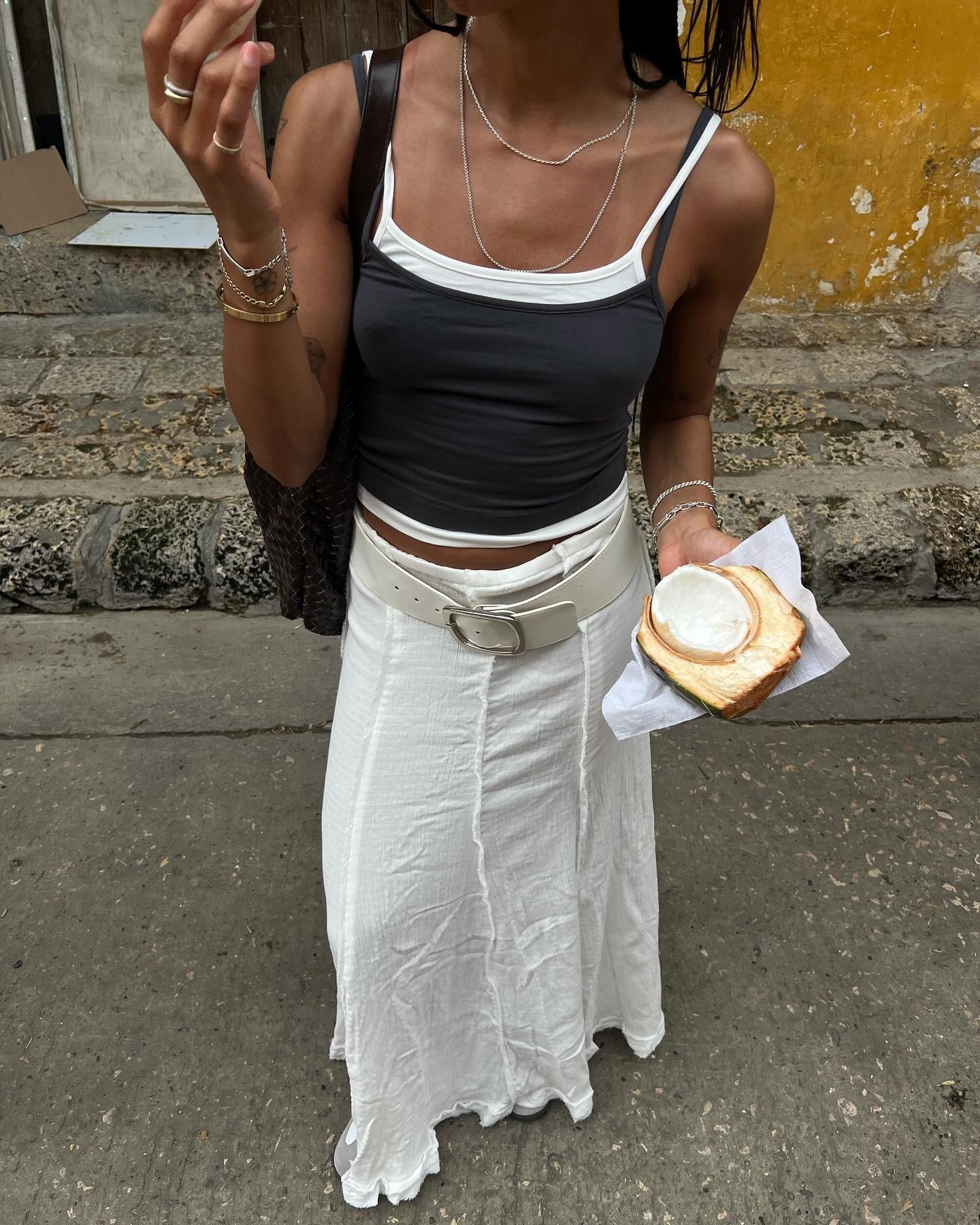 O look com regata apresenta uma combinação moderna e casual. A parte de cima é uma regata cinza com detalhes em branco, destacando-se por seu corte justo e contemporâneo. A saia longa branca, com textura leve, complementa o visual, enquanto um cinto largo