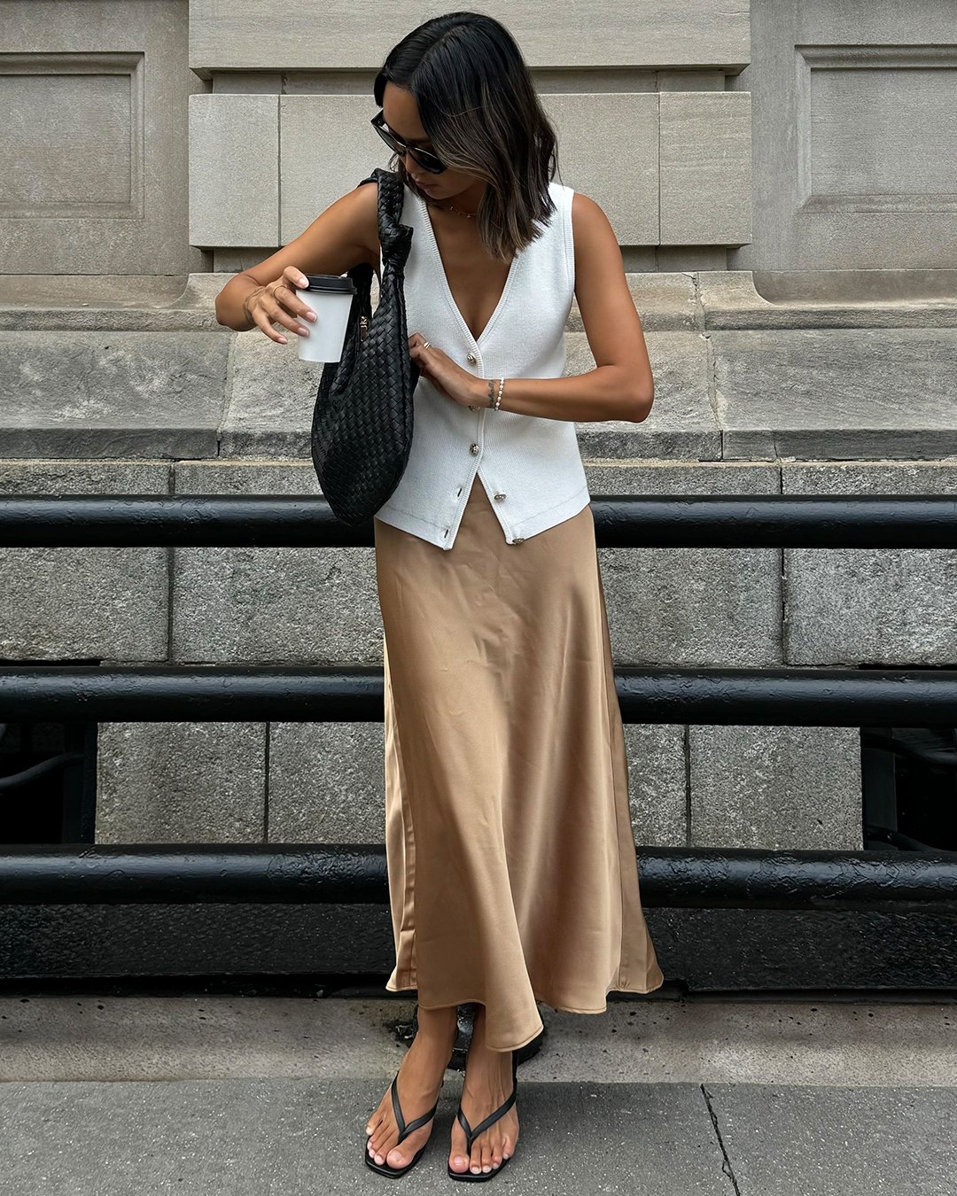 Mulher de colete branco e saia de cetim bege segura copo e bolsa.