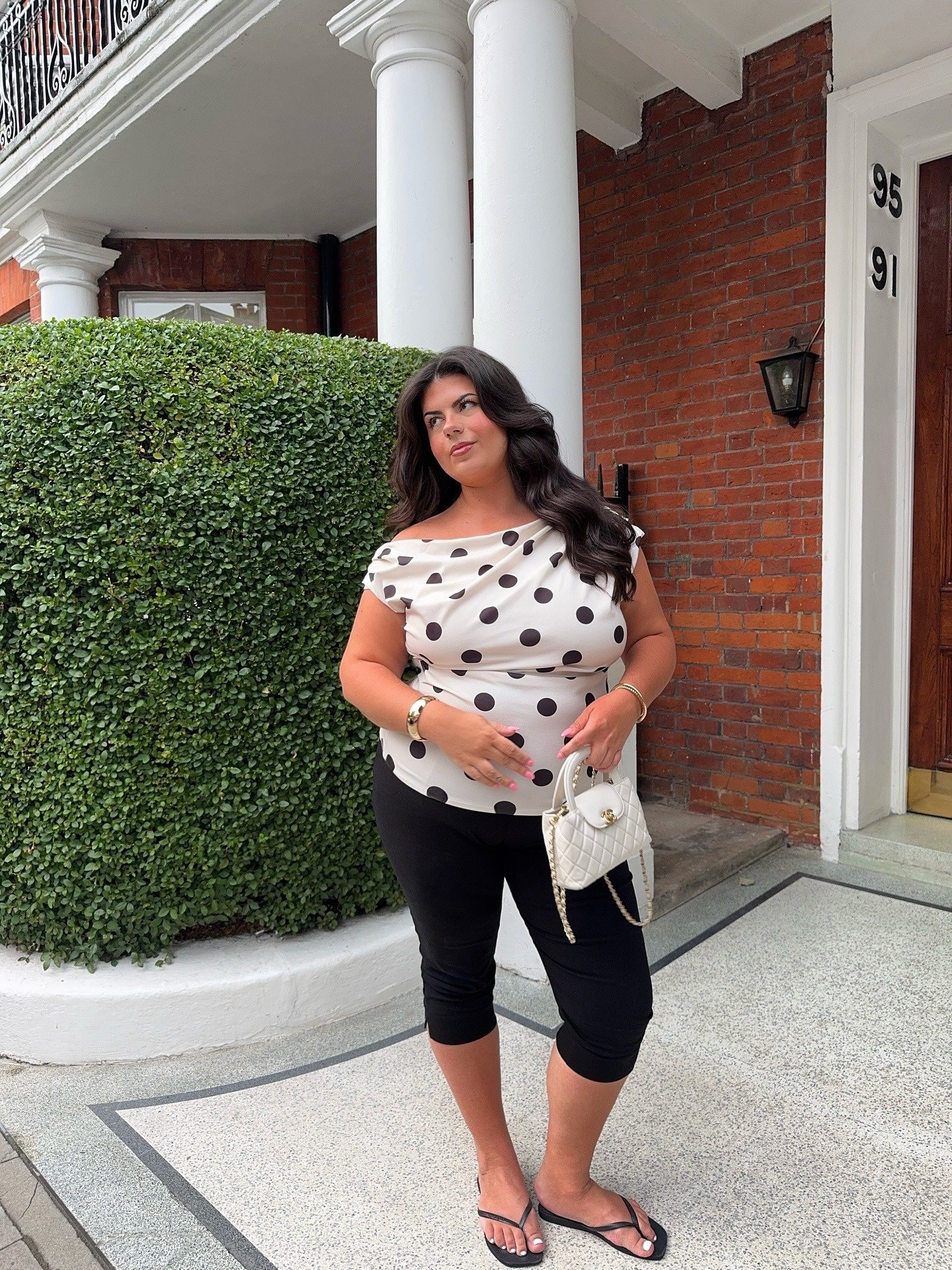 O look com sandália de dedo apresenta uma combinação elegante e casual. A blusa de bolinhas em tons neutros destaca-se sobre a calça preta, criando um visual equilibrado. A modelo segura uma bolsa branca, que complementa o estilo.