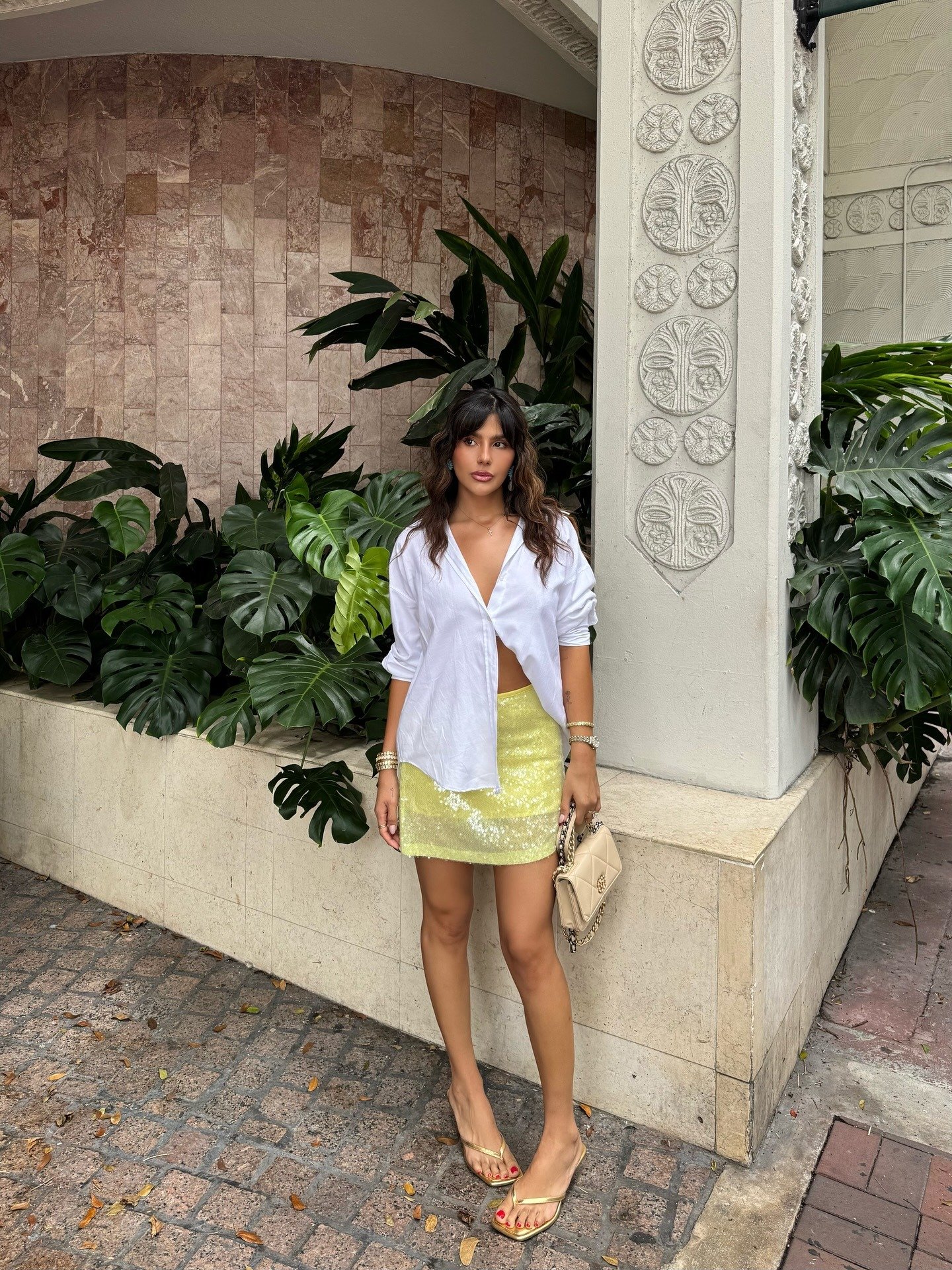 O look com sandália dourada destaca uma combinação vibrante e moderna. A blusa branca, de corte solto, contrasta harmoniosamente com a saia amarela de paetês, trazendo um toque de brilho. A sandália dourada, com design elegante, complementa o visual, enqu