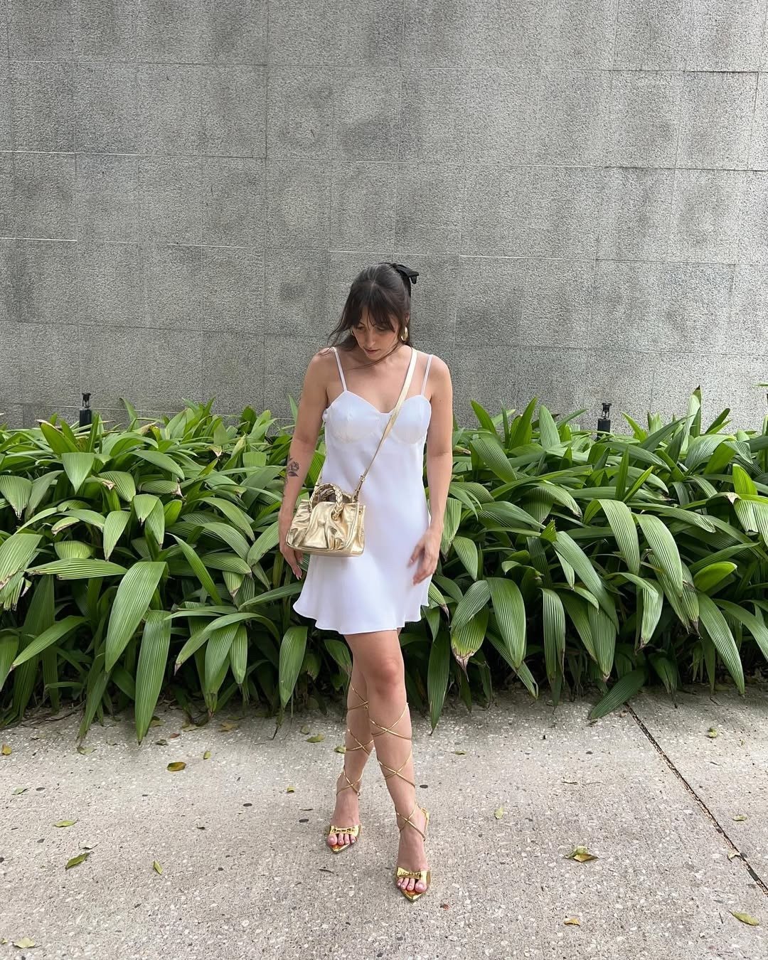 Look com sandália dourada destaca um vestido branco de alças finas, que exala leveza e sofisticação. A sandália, com tiras que sobem pelas pernas, adiciona um toque moderno e ousado ao visual. A bolsa dourada complementa o conjunto, criando harmonia nas t