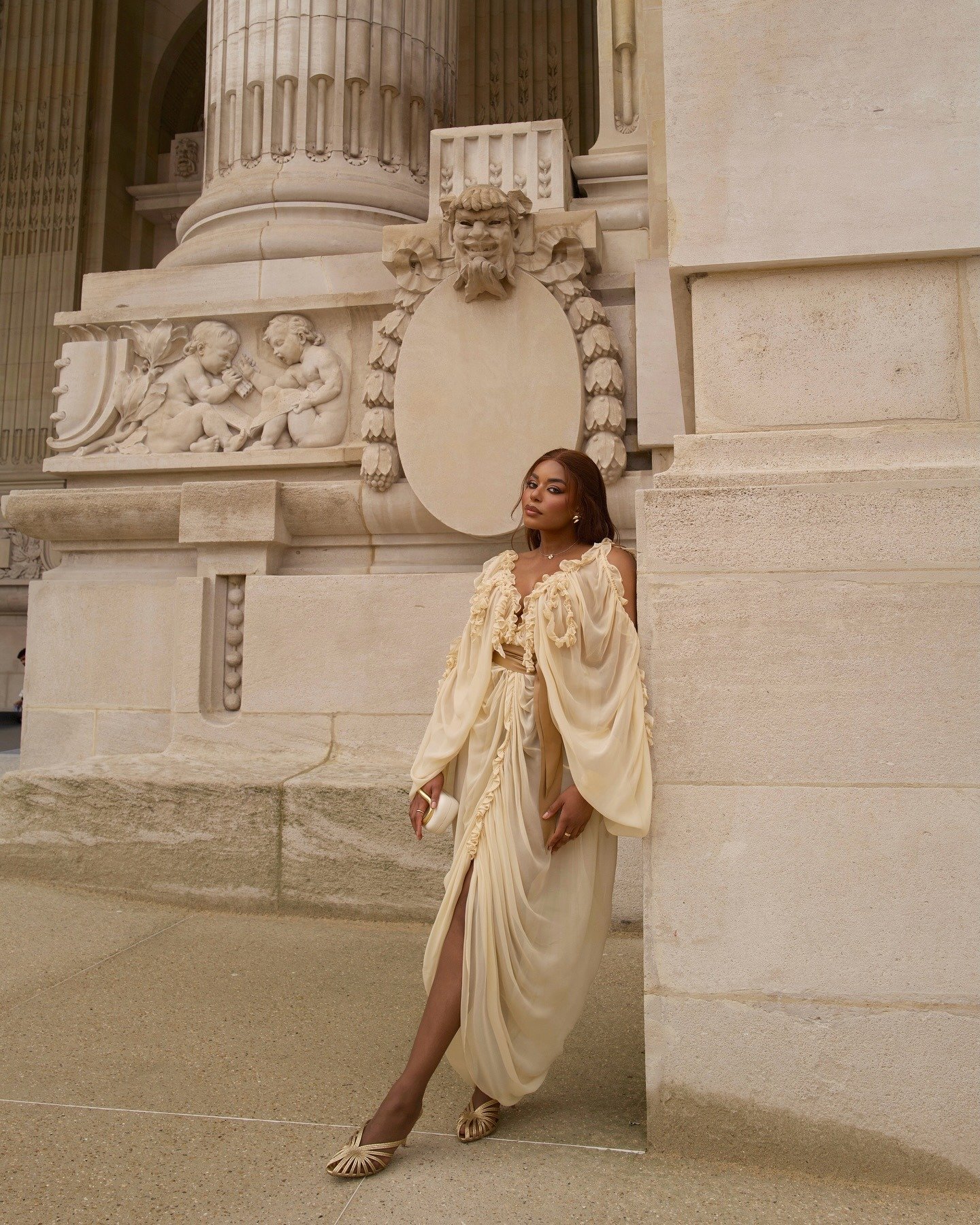 O look apresenta uma combinação sofisticada com uma sandália dourada que complementa a elegância do vestido em tom creme. O vestido, com detalhes fluidos e babados, exibe uma silhueta graciosa, enquanto a maquiagem sutil destaca o rosto. O cenário clássic