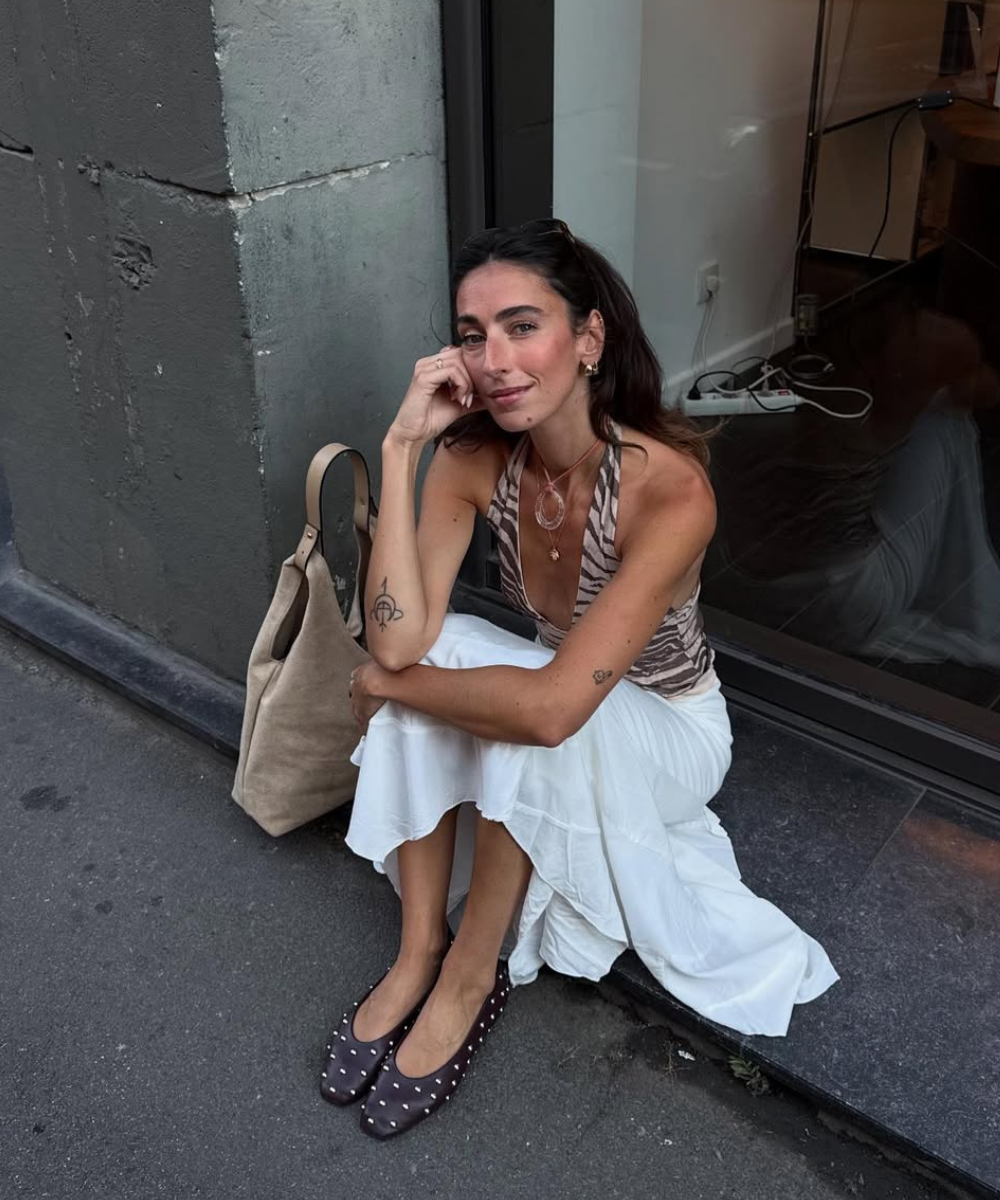 Mulher sentada na calçada com saia branca, blusa estampada e sapatilha de tachas. Bolsa ao lado.