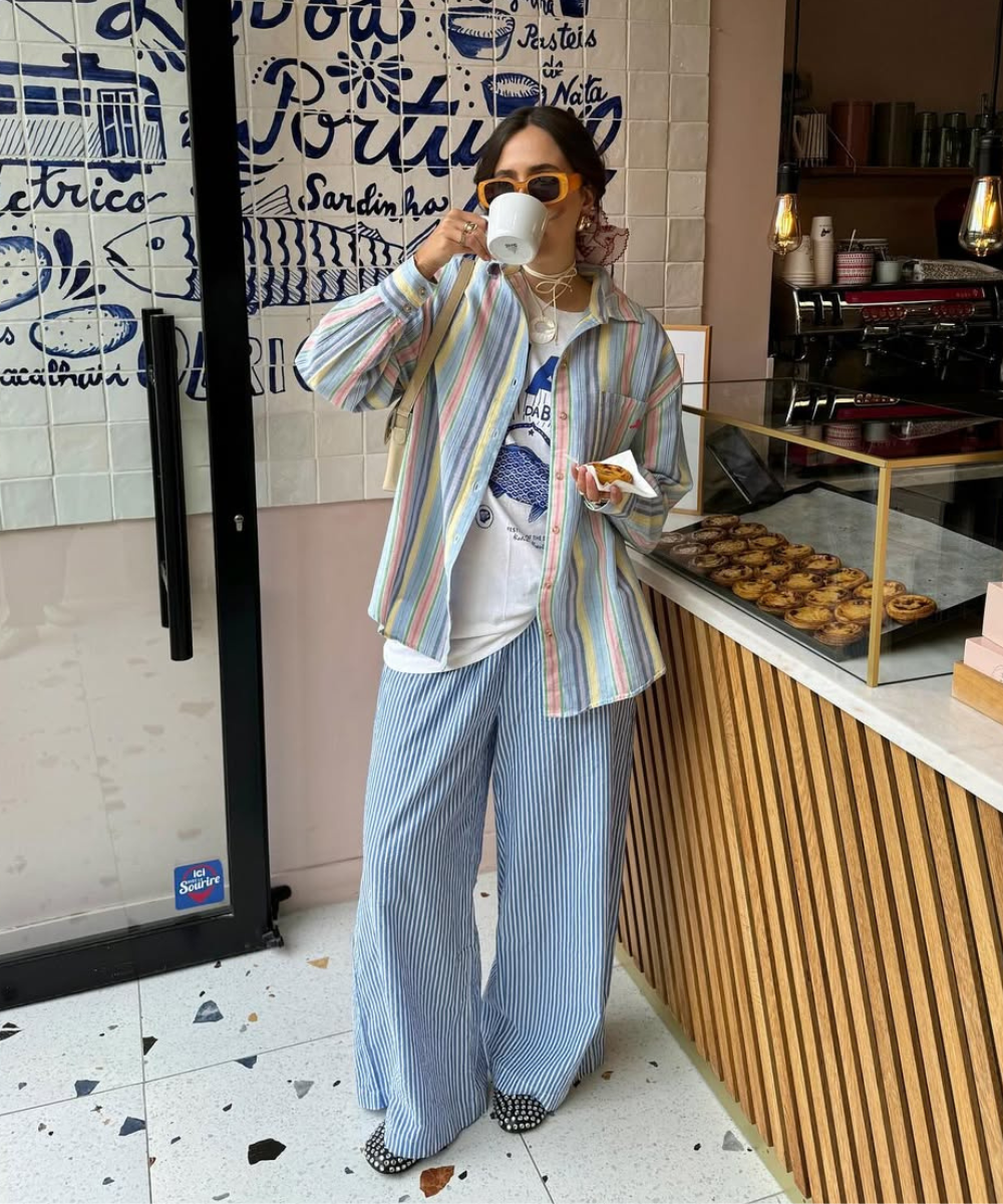Pessoa com xícara de café, usa roupa listrada colorida e sapatilha de tachas, em padaria com pastéis de nata.