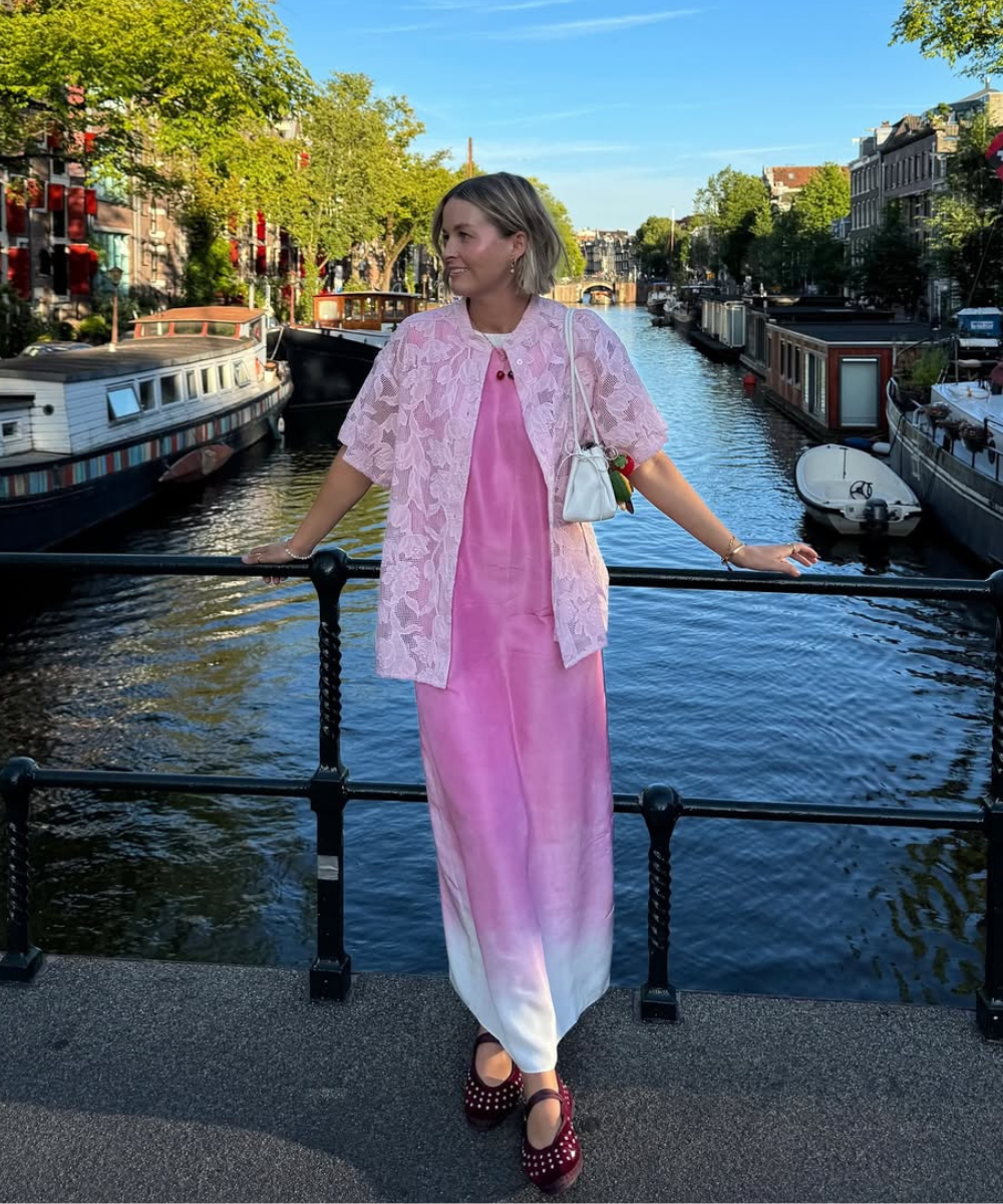 Mulher de vestido rosa posa em ponte, usando sapatilha de tachas, com canal e barcos ao fundo.