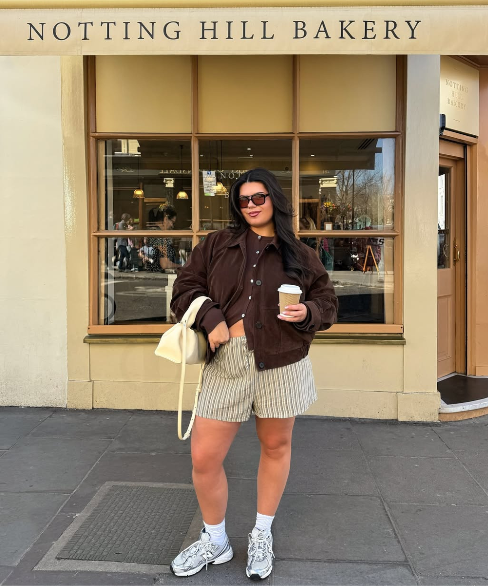 Pessoa em frente à padaria, usando jaqueta marrom e shorts listrado, segurando café e bolsa clara.