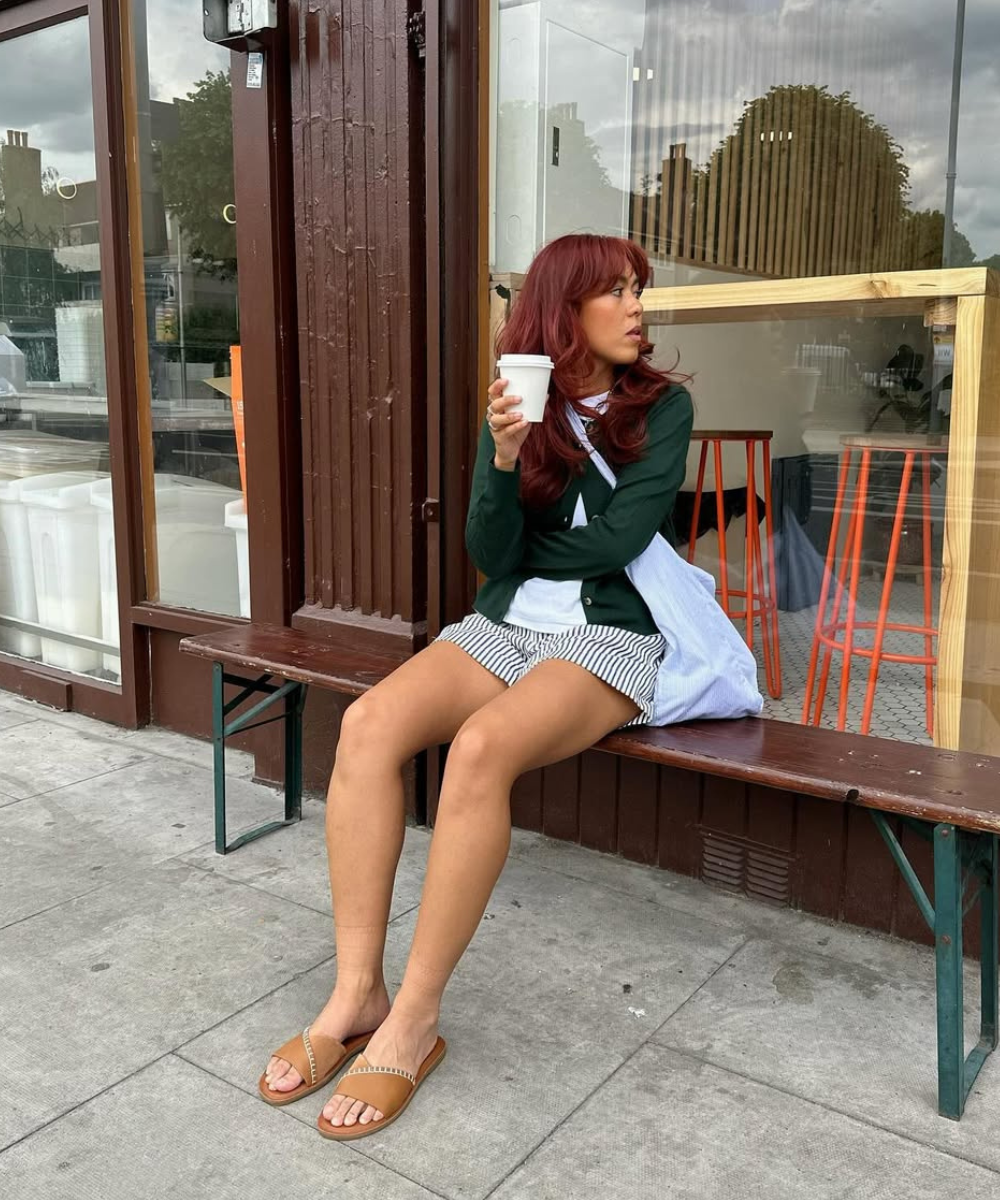Pessoa sentada em um banco segurando um copo de café, vestindo shorts listrado e suéter verde.