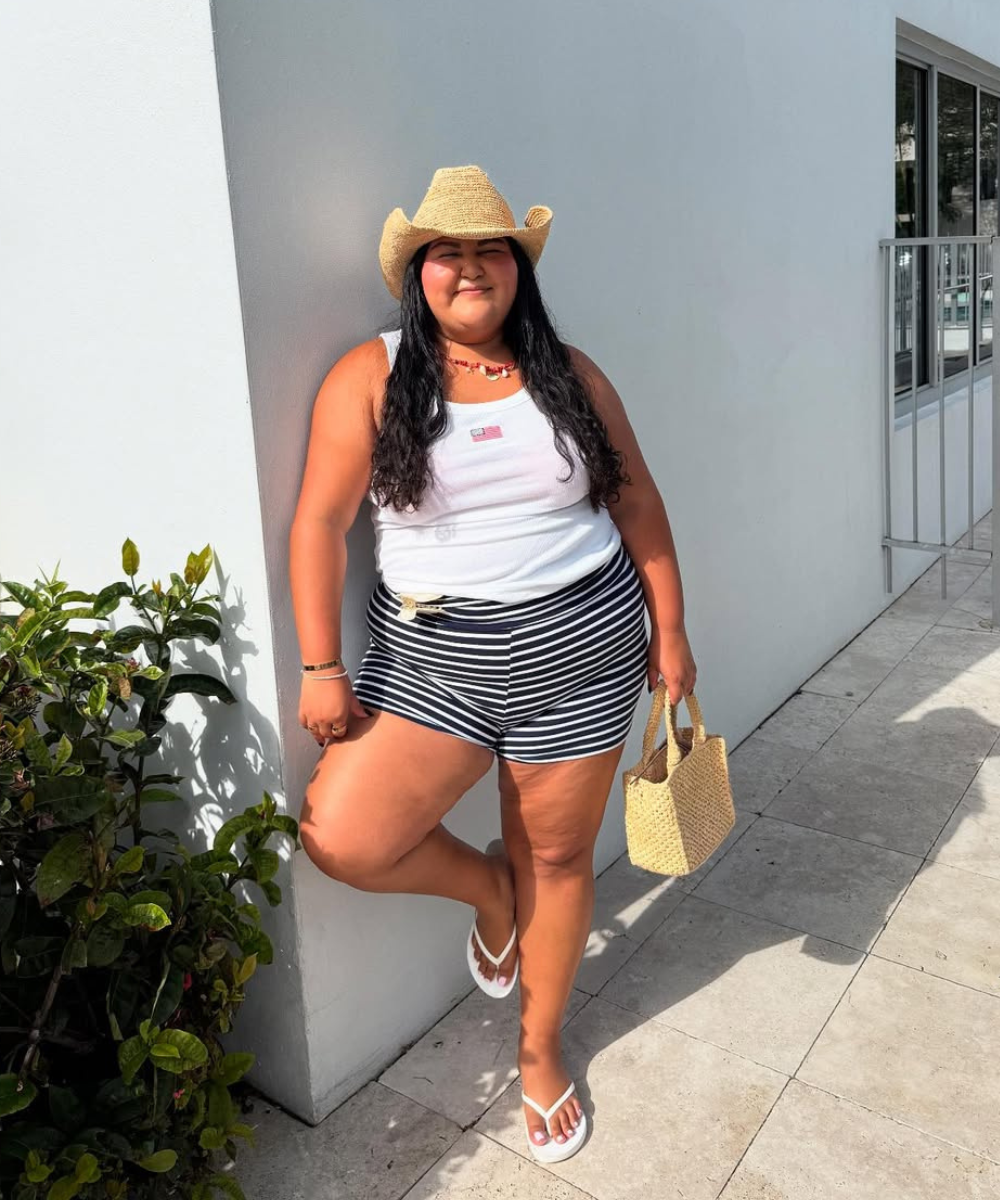 Mulher com chapéu de palha e shorts listrado preto e branco, apoiada em parede branca, segurando bolsa de palha.