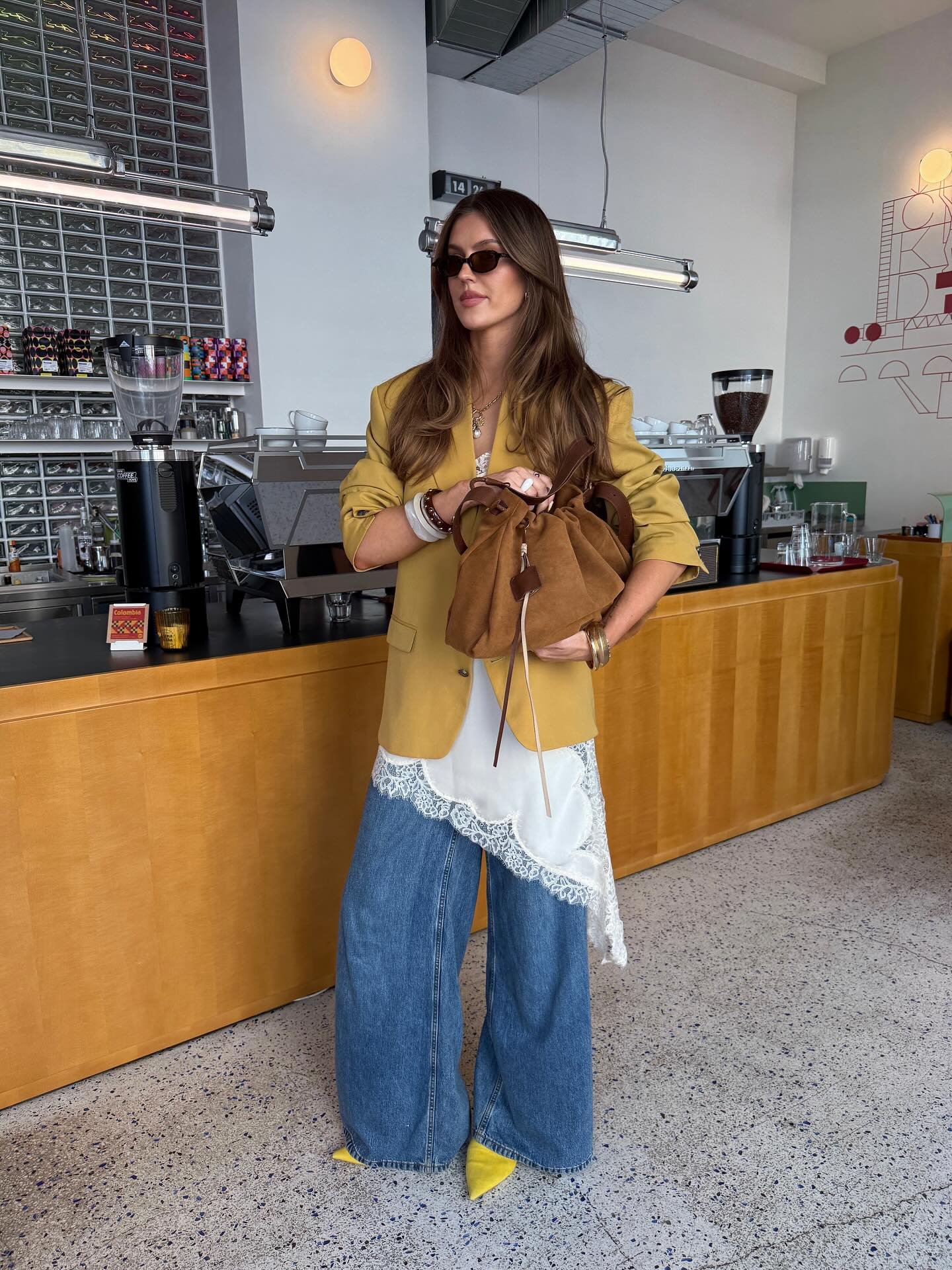 Casaco amarelo sobre slip dress assimétrico com detalhe de renda, combinado com calça jeans e sapatos amarelos. A pessoa segura uma bolsa marrom em um ambiente moderno, possivelmente um café.