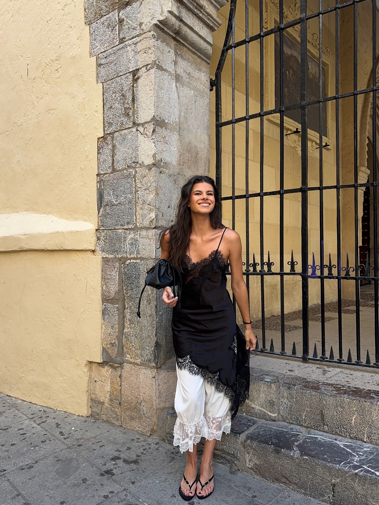 Mulher sorridente usa slip dress assimétrico preto e branco com renda, em frente a um prédio antigo.