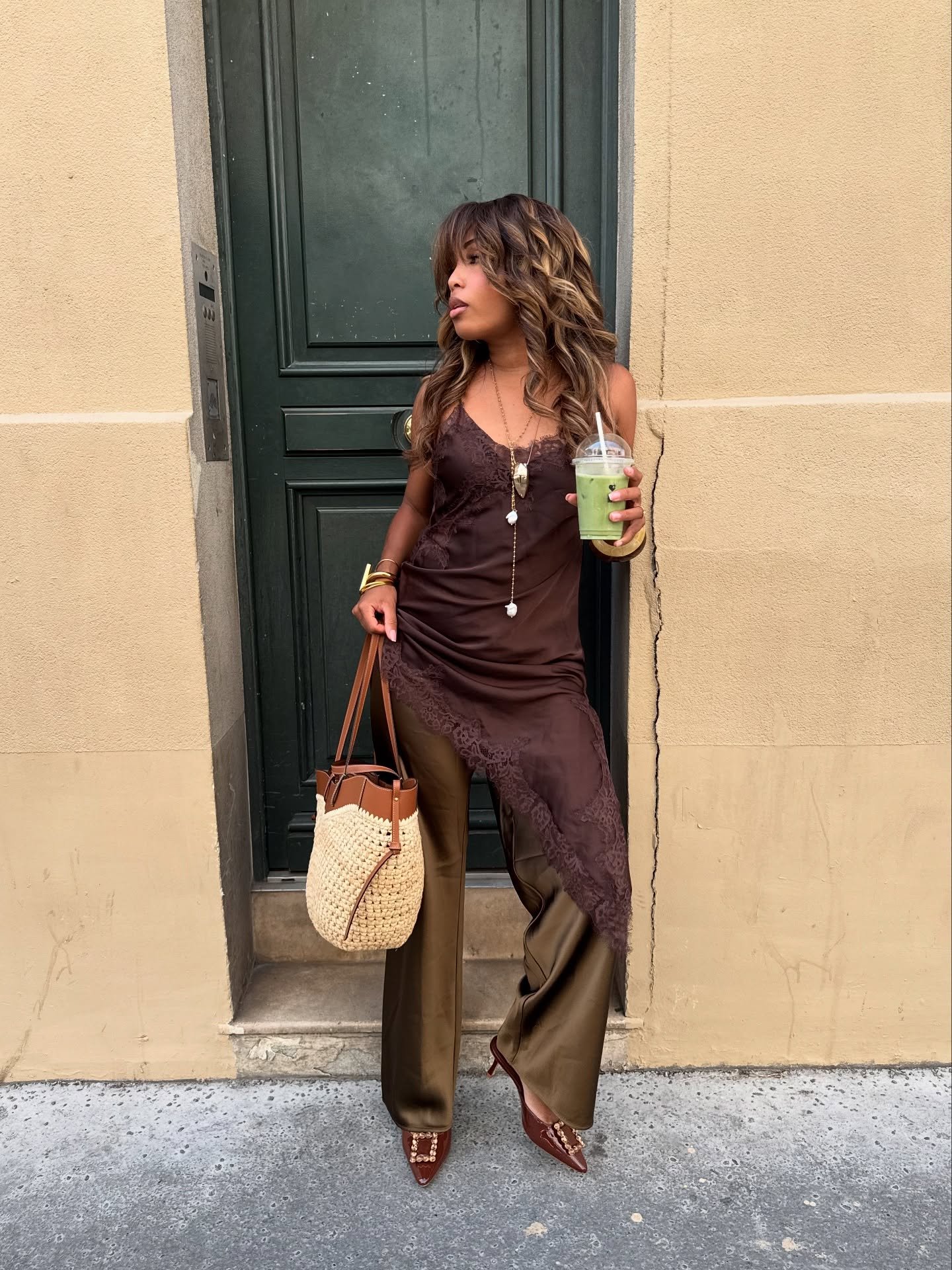 Mulher com slip dress assimétrico marrom sobre calça verde-oliva, segurando bebida verde, ao lado de porta verde escura.