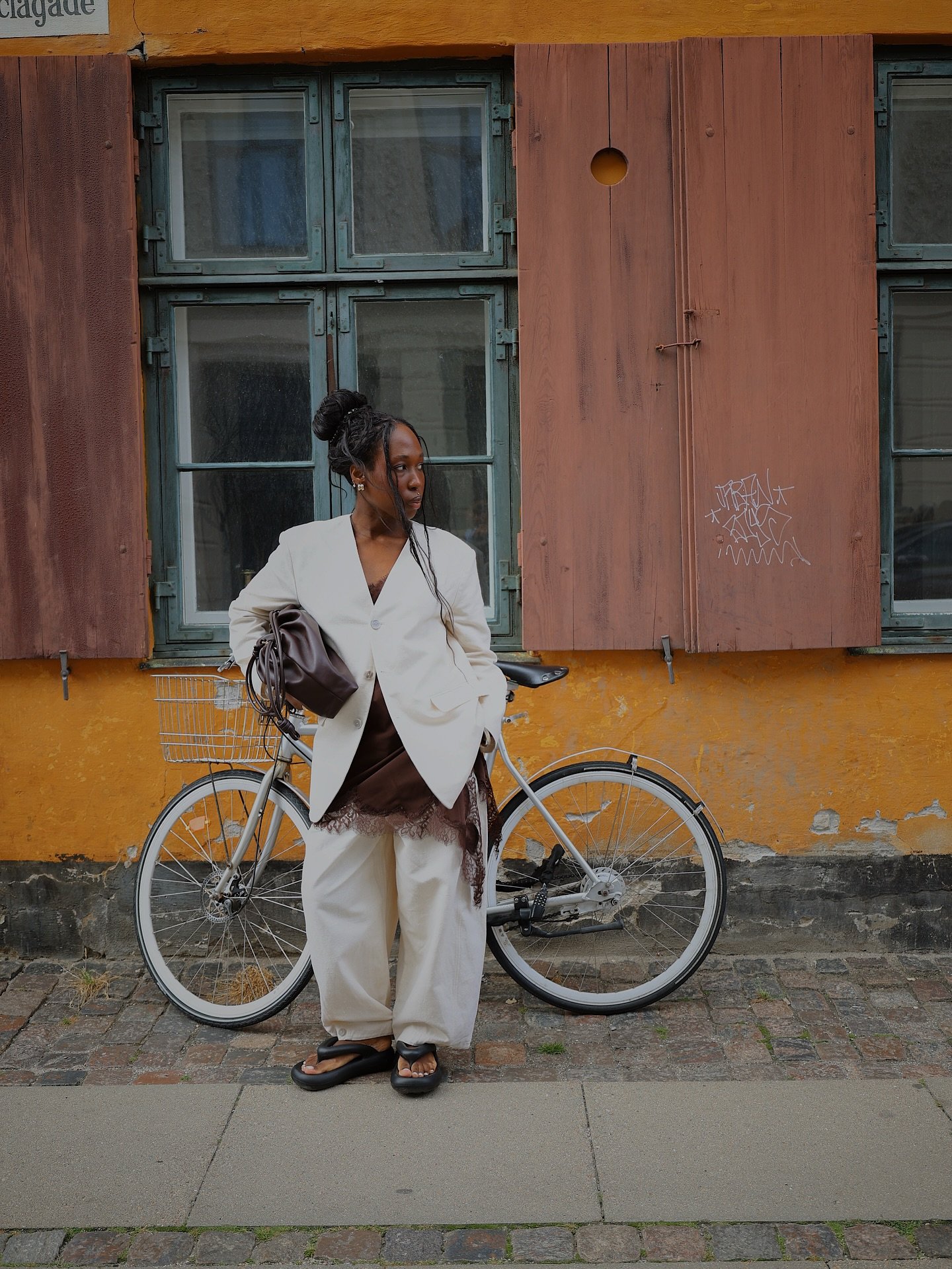 Pessoa em pé ao lado de uma bicicleta, veste um blazer claro sobre slip dress assimétrico e calça larga.
