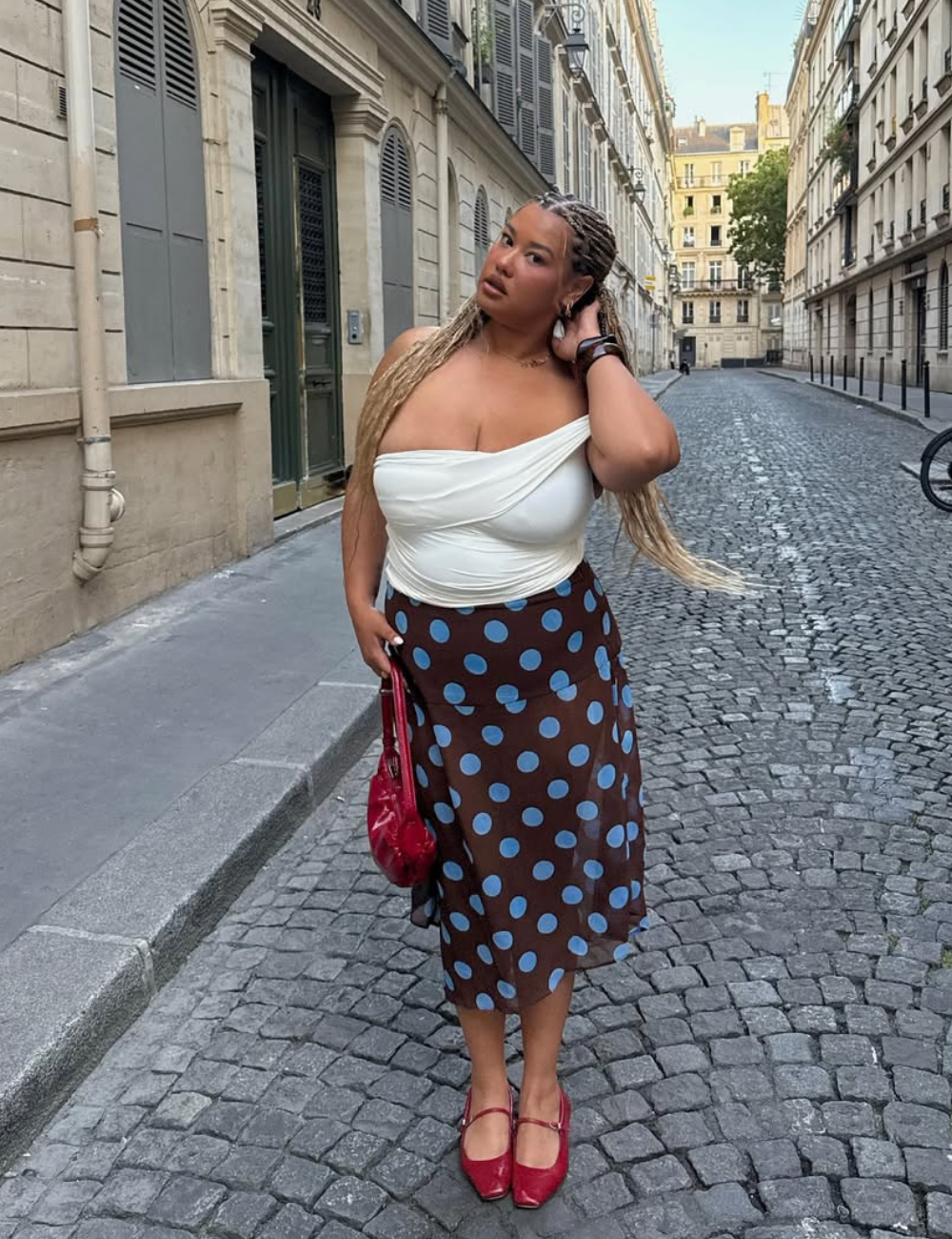 Nesta cena urbana, uma mulher exibe um look estiloso com saia, combinando uma blusa branca ajustada e uma saia midi marrom com bolinhas azuis. O visual é complementado por sapatos vermelhos e uma bolsa na mesma tonalidade. O cenário parisiense, com suas r