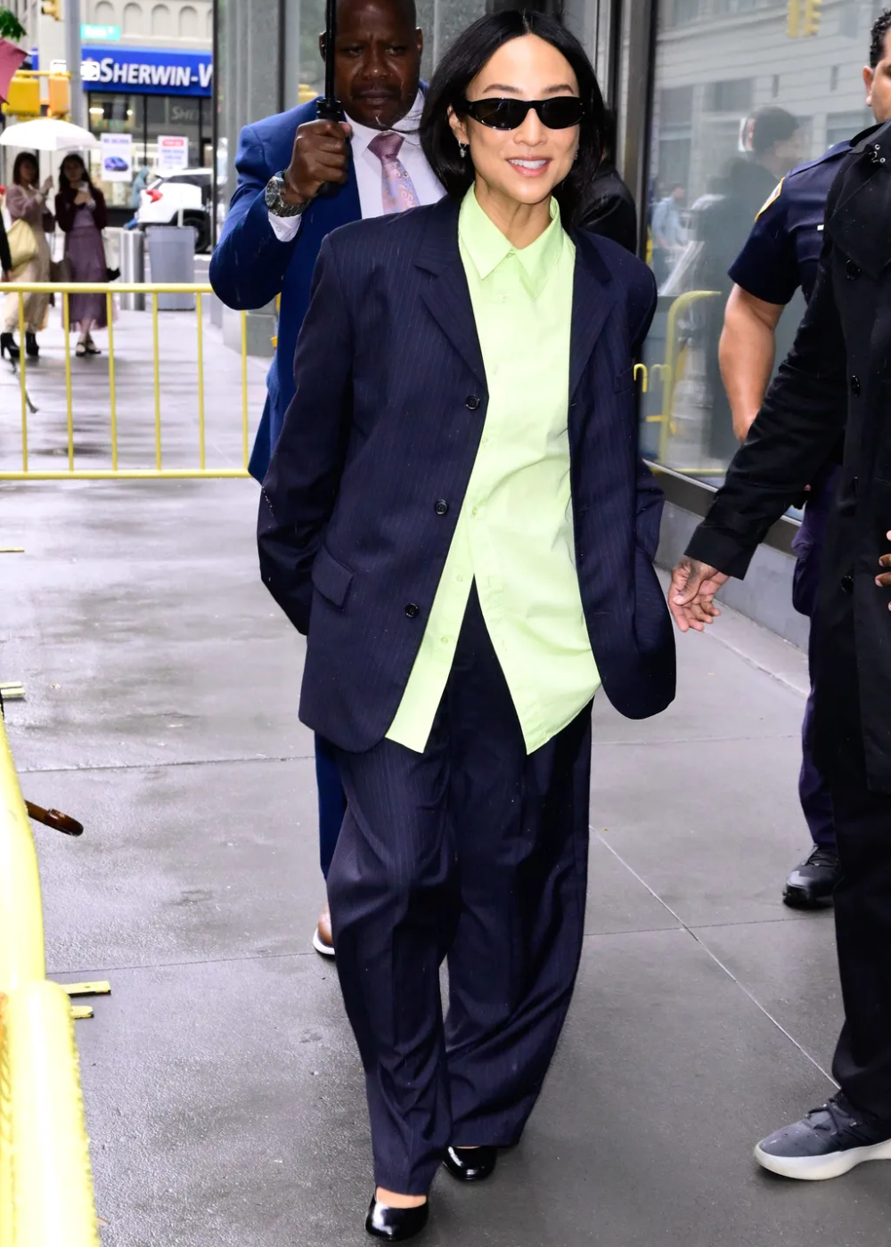 O look apresenta um blazer listrado em azul escuro, combinando com uma camisa verde-clara. A modelagem oversized do blazer traz um ar contemporâneo e descontraído. O uso de óculos escuros complementa o visual, conferindo sofisticação.