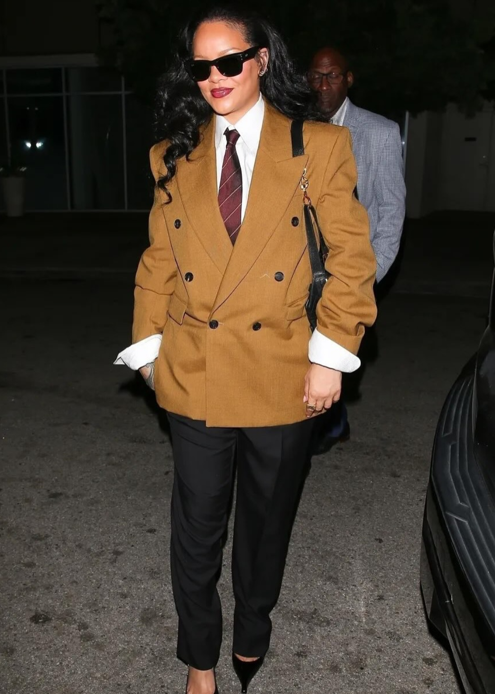 O look apresenta um blazer oversized em tom mostarda, combinando com uma camisa branca e uma gravata marrom. As calças pretas ajustadas e os sapatos de salto alto completam o visual elegante. Óculos escuros e cabelo solto adicionam um toque moderno.