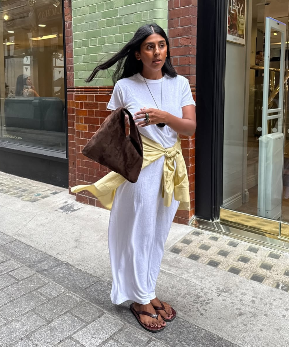 Pessoa caminhando na calçada usa vestido branco, jaqueta na cintura e bolsa marrom. Visual leve e despojado.