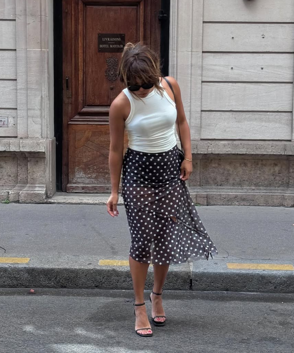 Mulher com regata branca e saia preta de bolinhas, em calçada urbana. Visual leve e moderno. Looks fresquinhos com branco.