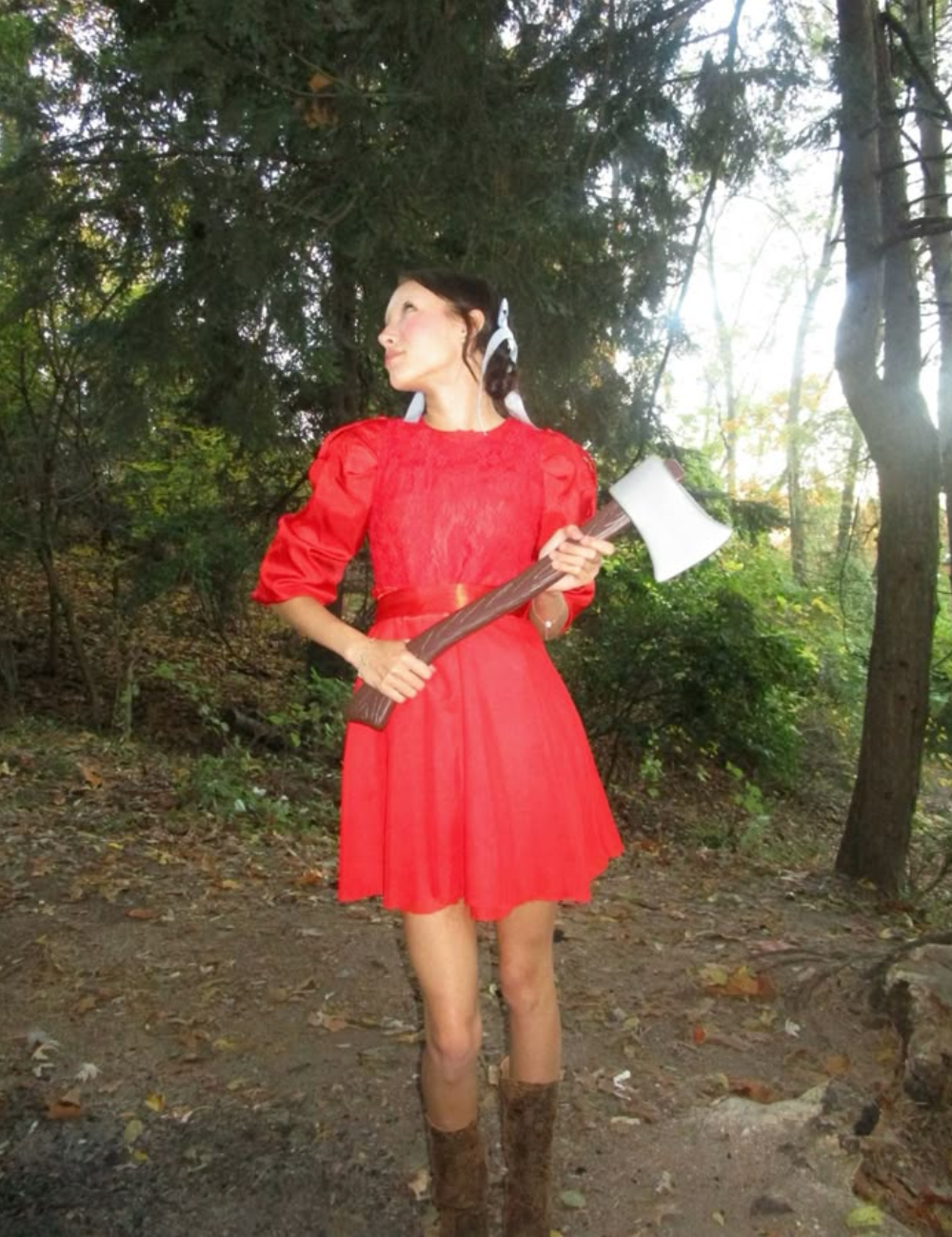 Uma jovem usa uma fantasia de Halloween simples, inspirada em contos de fadas, com um vestido vermelho vibrante e mangas bufantes. Ela segura um machado de brinquedo, criando um contraste interessante entre a inocência da roupa e o acessório ameaçador.